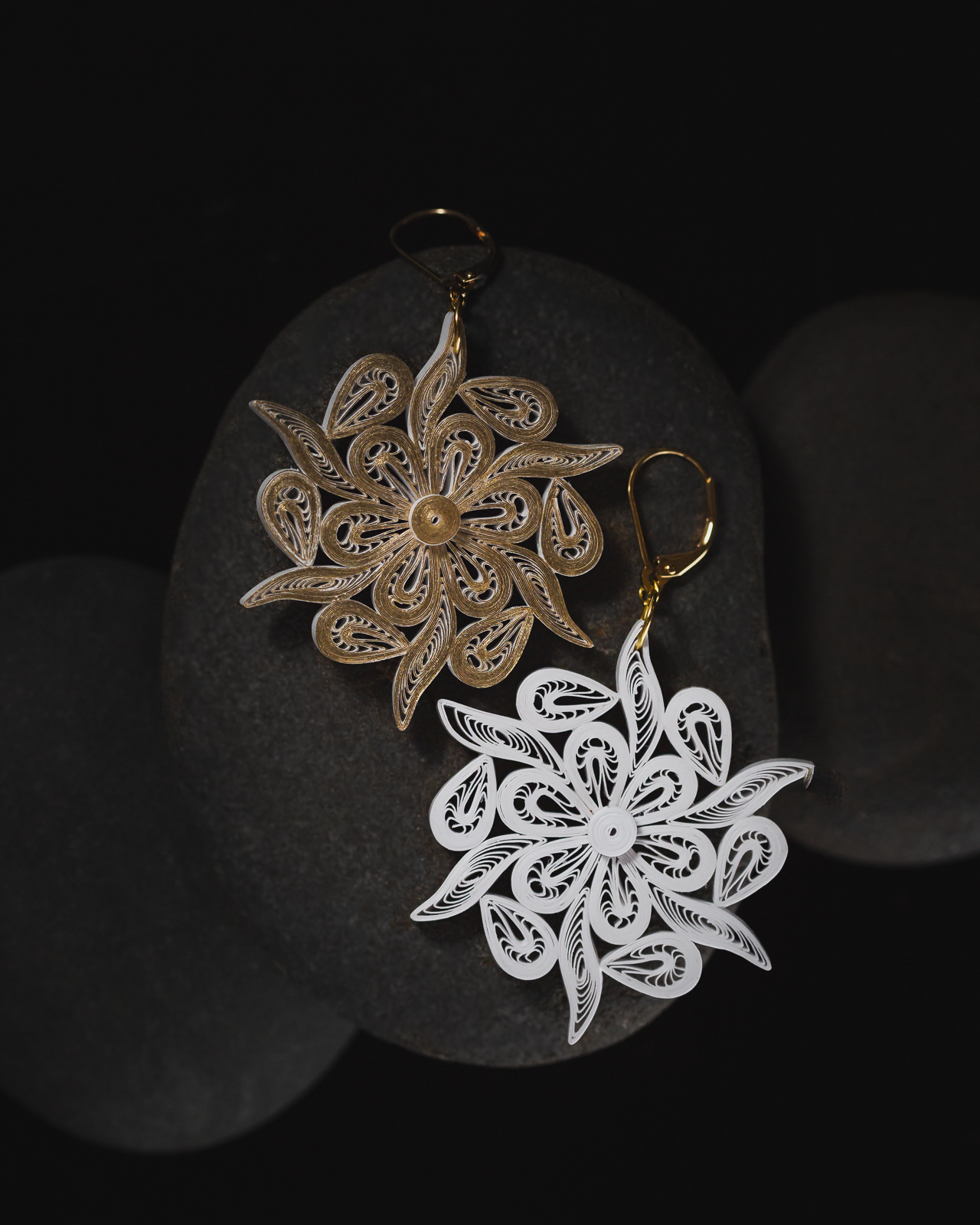 Two intricate snowflake-shaped earrings, one gold and one white, resting on dark stones, with a black background.
