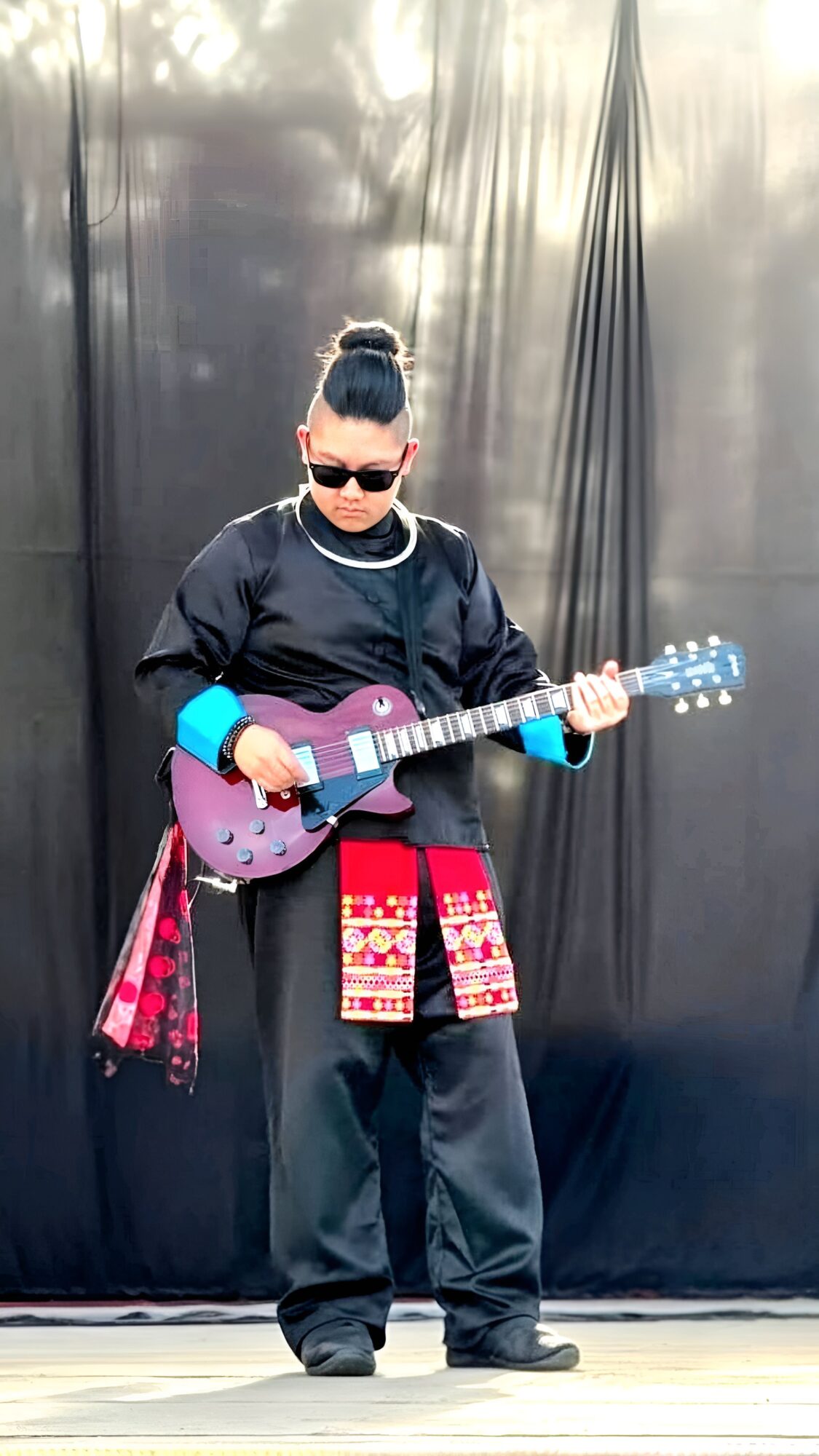 Person with a bun hairstyle, sunglasses, black jacket, and patterned skirt playing a purple guitar on stage.