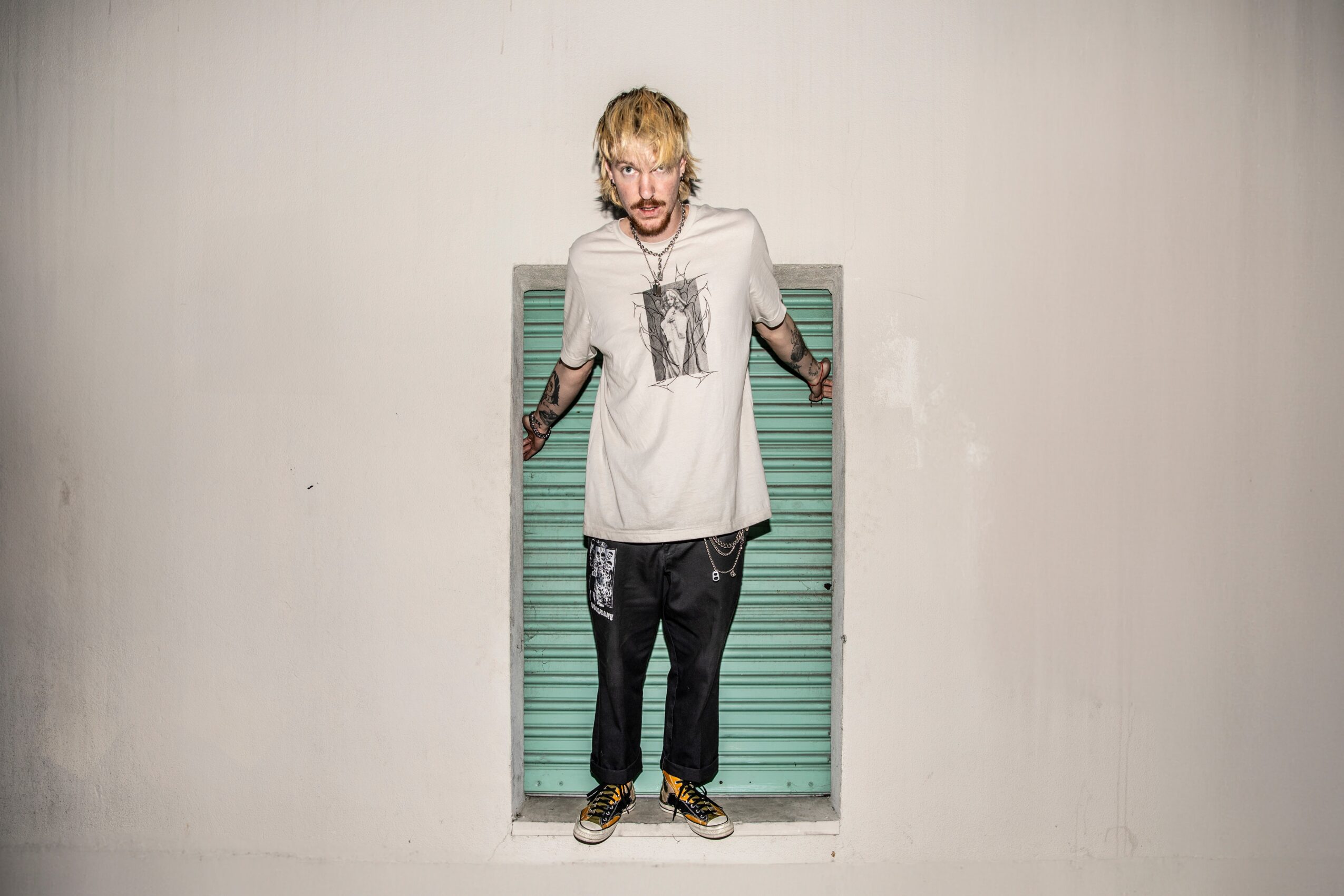 Person with blond hair and glasses standing in front of a teal window shutter, wearing a white t-shirt and black pants.