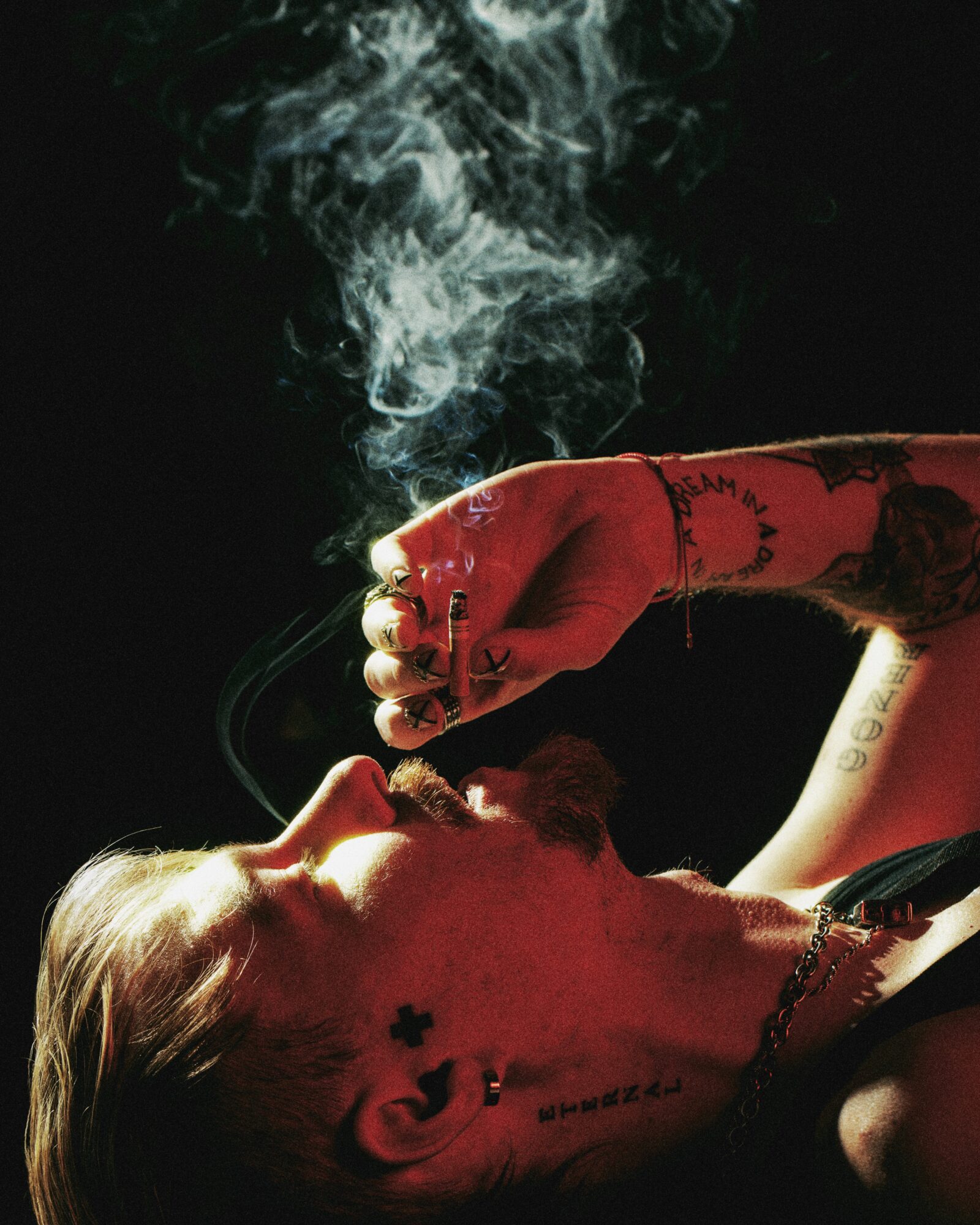 Person with tattoos smoking, smoke rising, dark background, side profile, red clothing, visible tattoos on arm and neck.