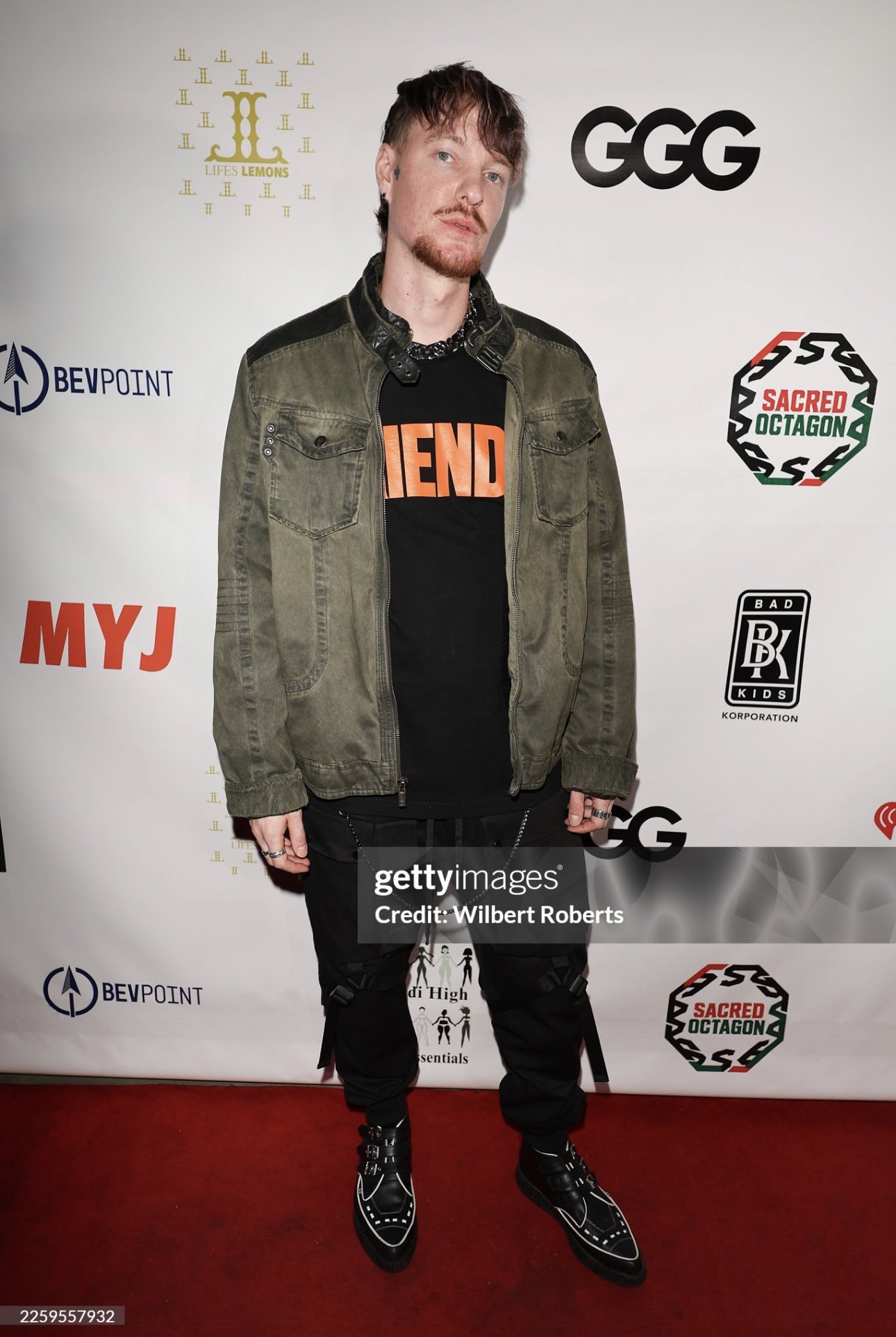 Young man with short hair and beard standing on red carpet, wearing a green jacket, black hoodie, and black shoes.