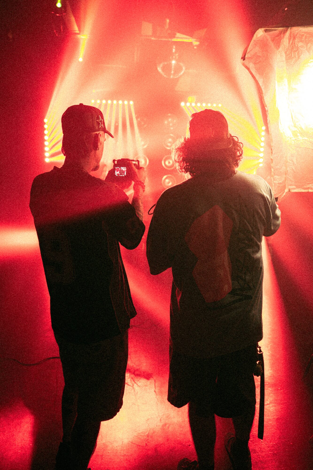 Two people stand in front of a stage with bright red and yellow lights, one taking a photo, silhouettes visible.