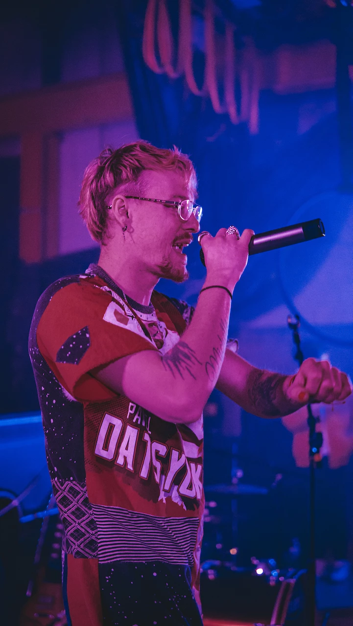 Person singing into a microphone on stage with colorful lighting, wearing glasses and a patterned shirt.