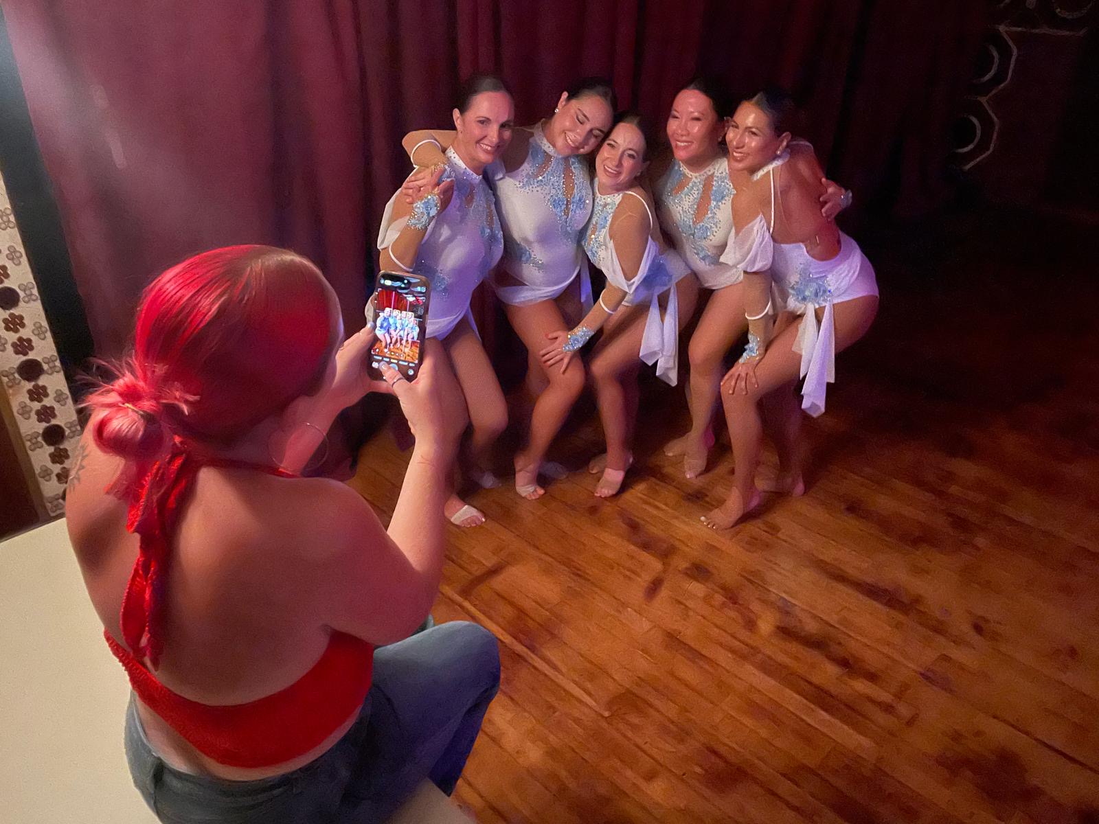 Group of five women in costumes posing for a photo, with a woman with red hair taking the picture, on a wooden floor.