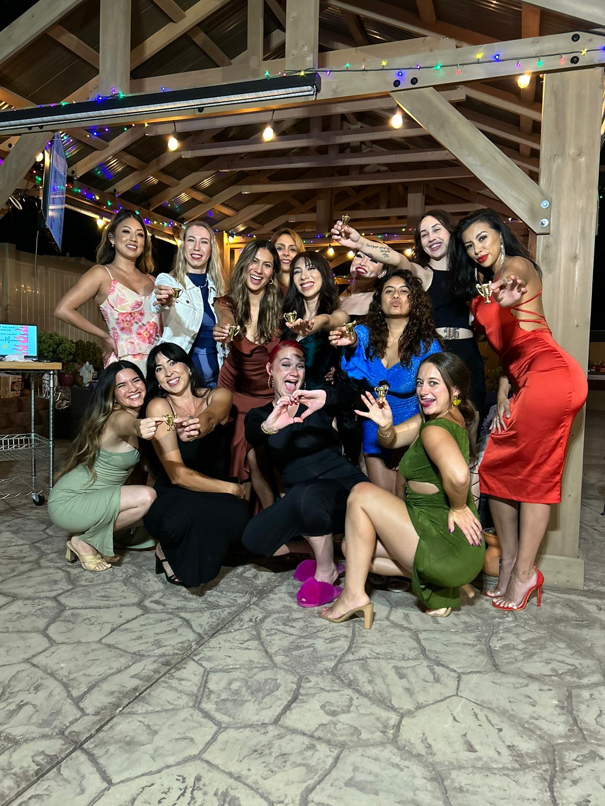 Group of women posing together indoors, some making peace signs, in colorful dresses, with wooden beams and string lights above.