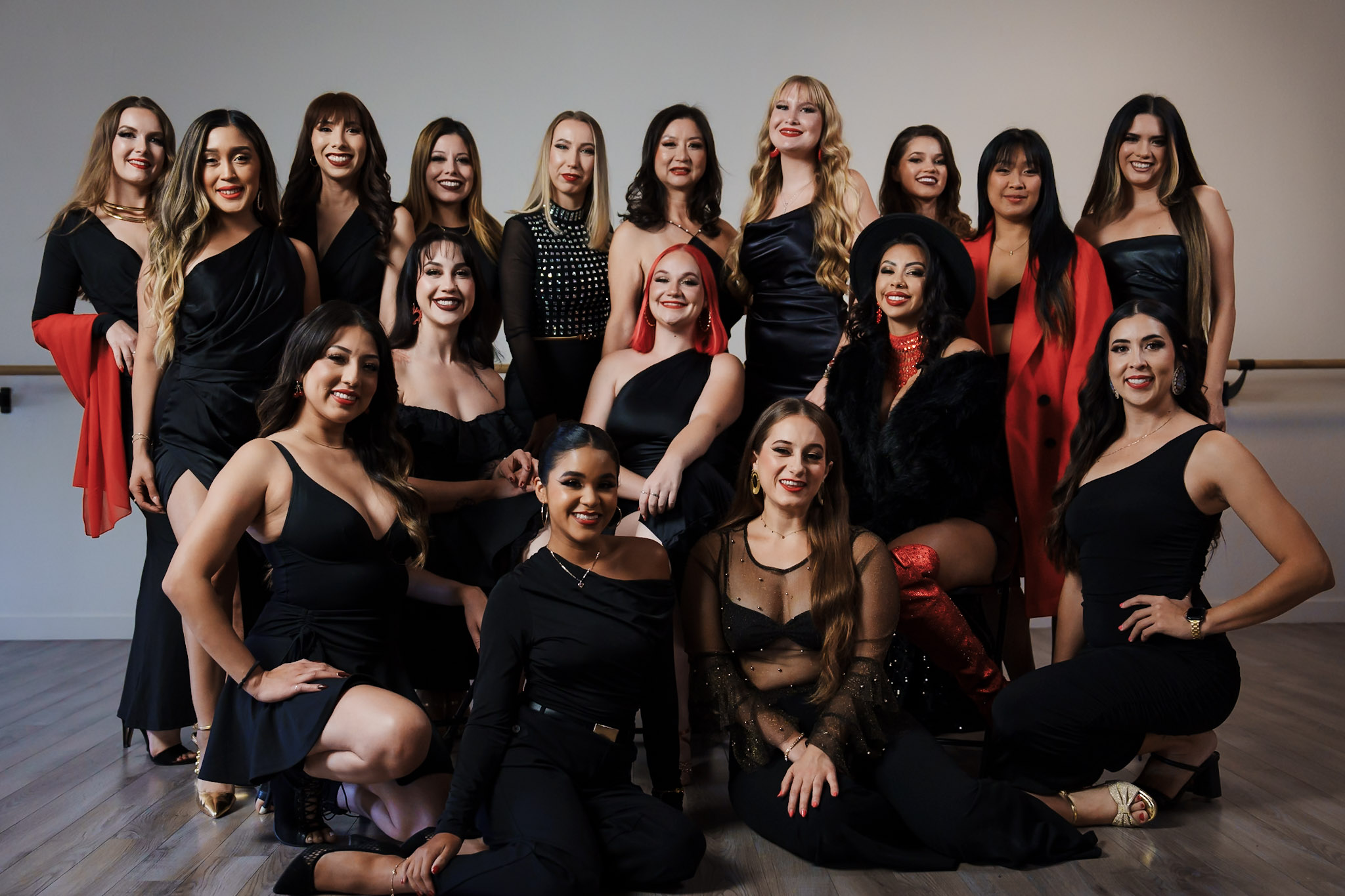 Group of 16 women in black dresses posing together indoors, some with red accents, smiling and standing or sitting.