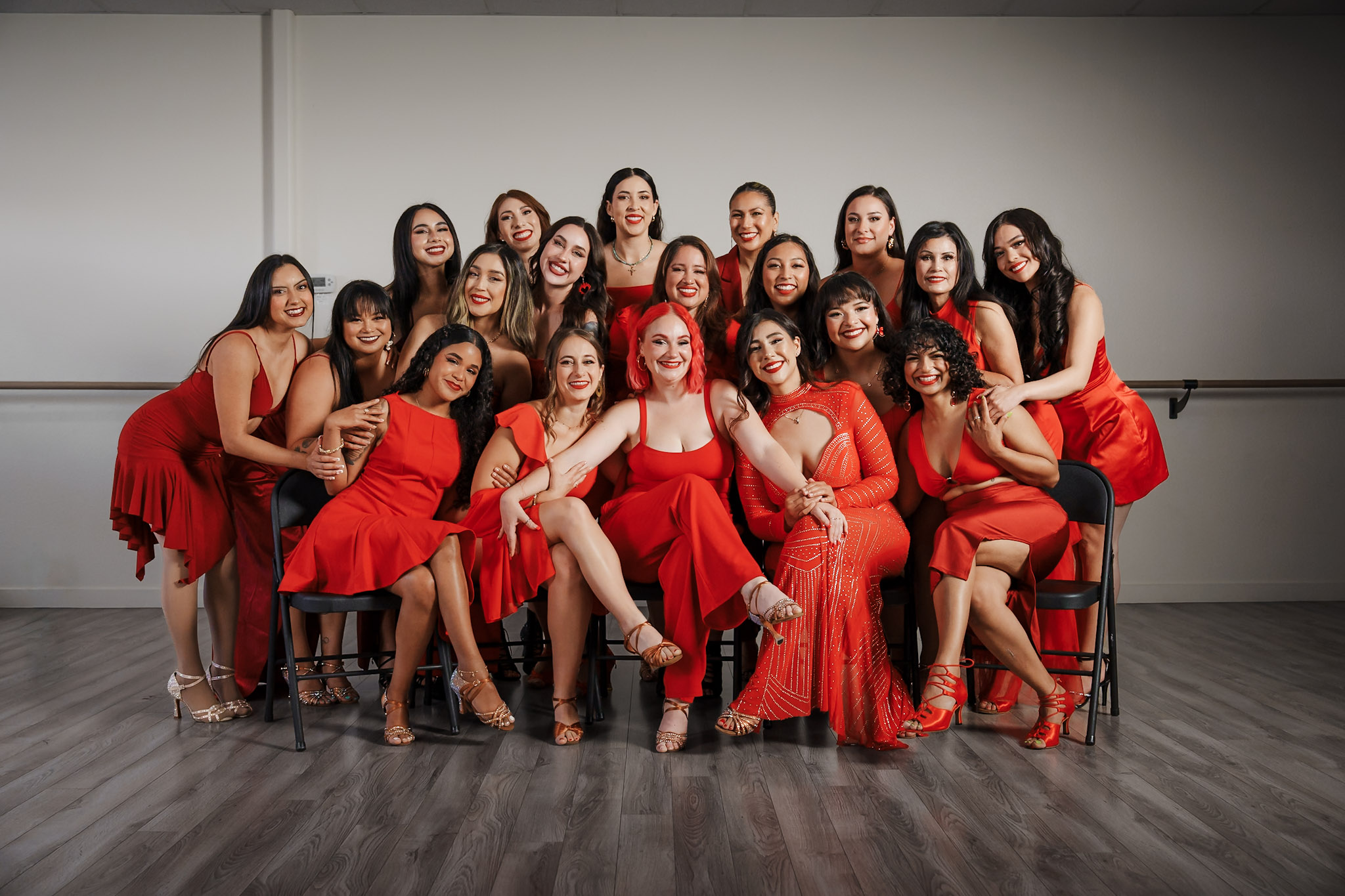 Group of women in red dresses sitting and standing in a room with a plain wall, smiling and posing together.