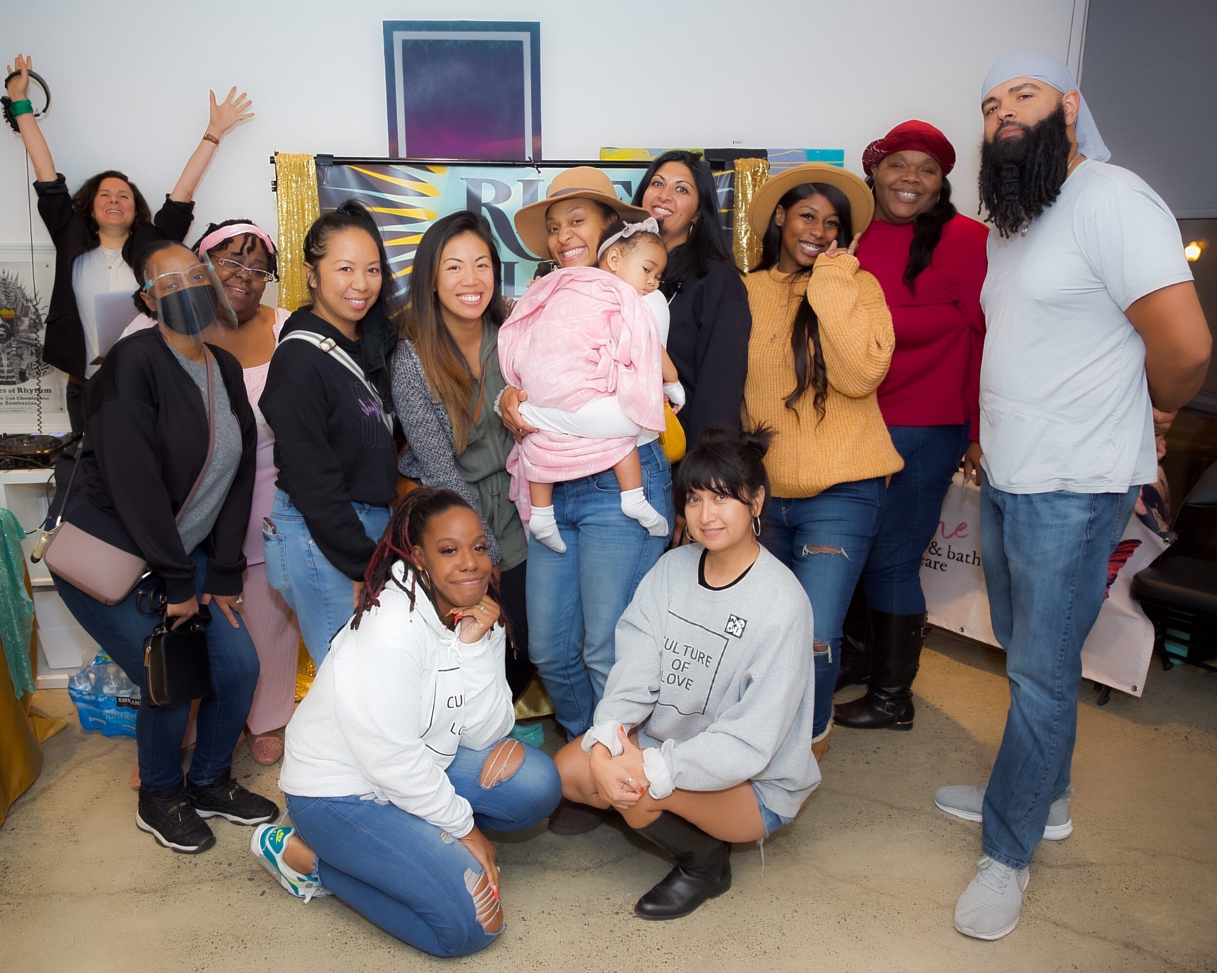 Group of diverse people smiling indoors, some standing and some kneeling, with colorful artwork in background.