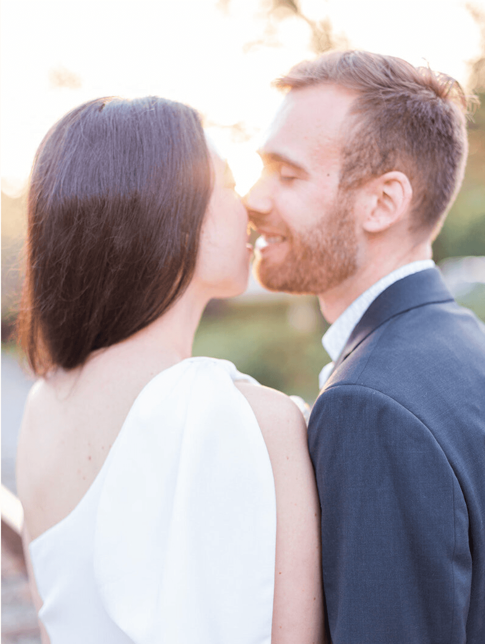 A couple faces each other outdoors, close and smiling, with sunlight behind them, in a romantic moment.