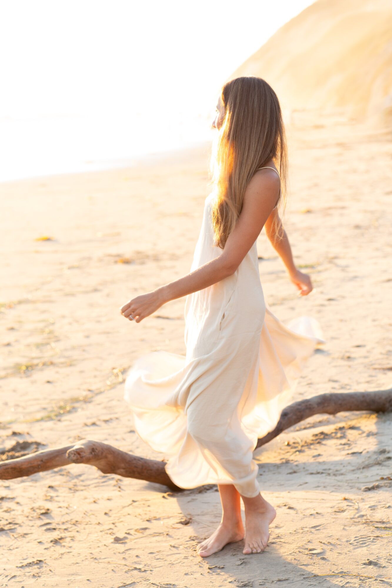 Woman in a flowing white dress balancing on a log on sandy terrain, with a large sand dune in the background.