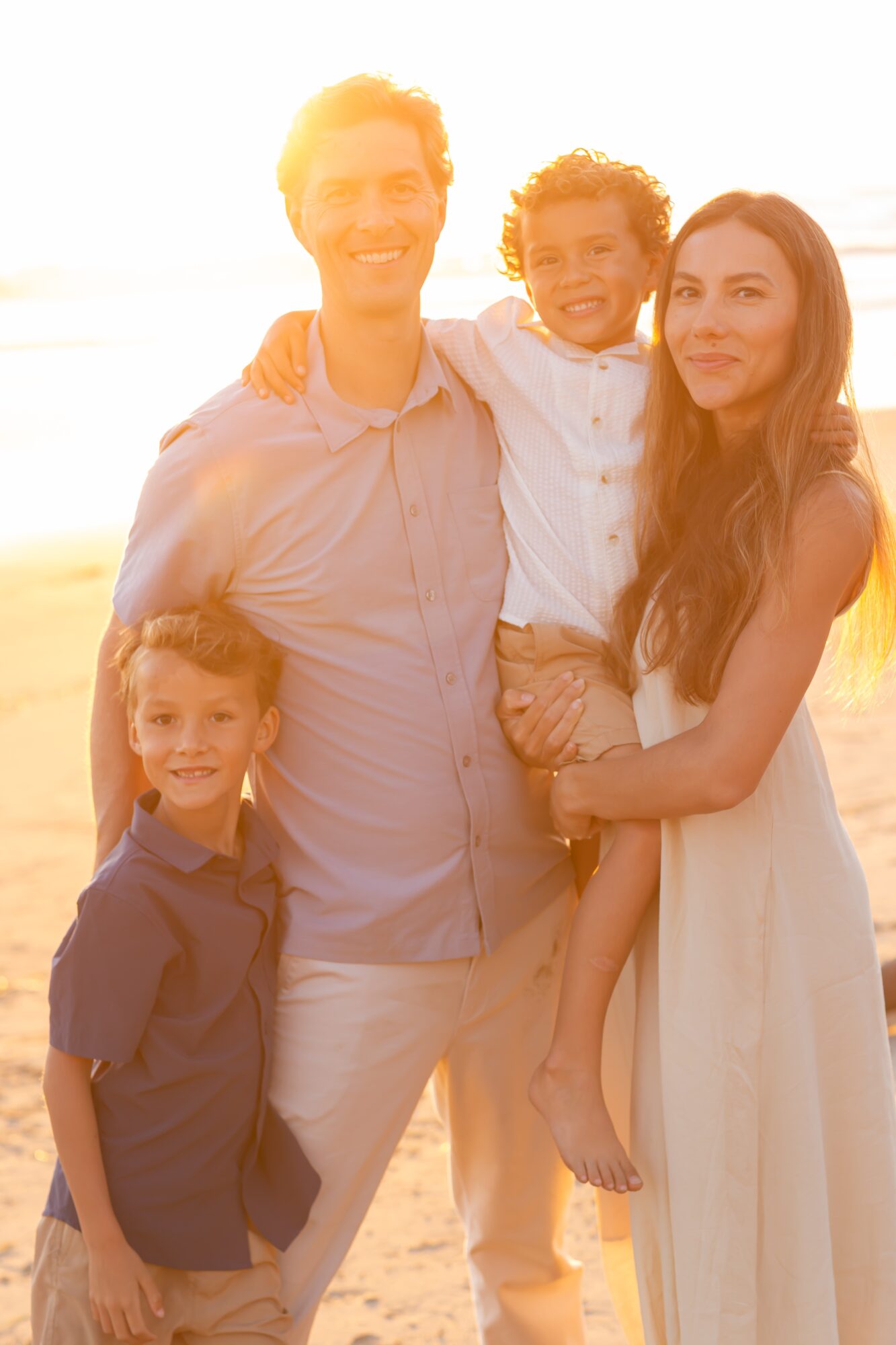 Family of four smiling outdoors at sunset, with a man, woman, and two children, one girl and one boy, on a beach.