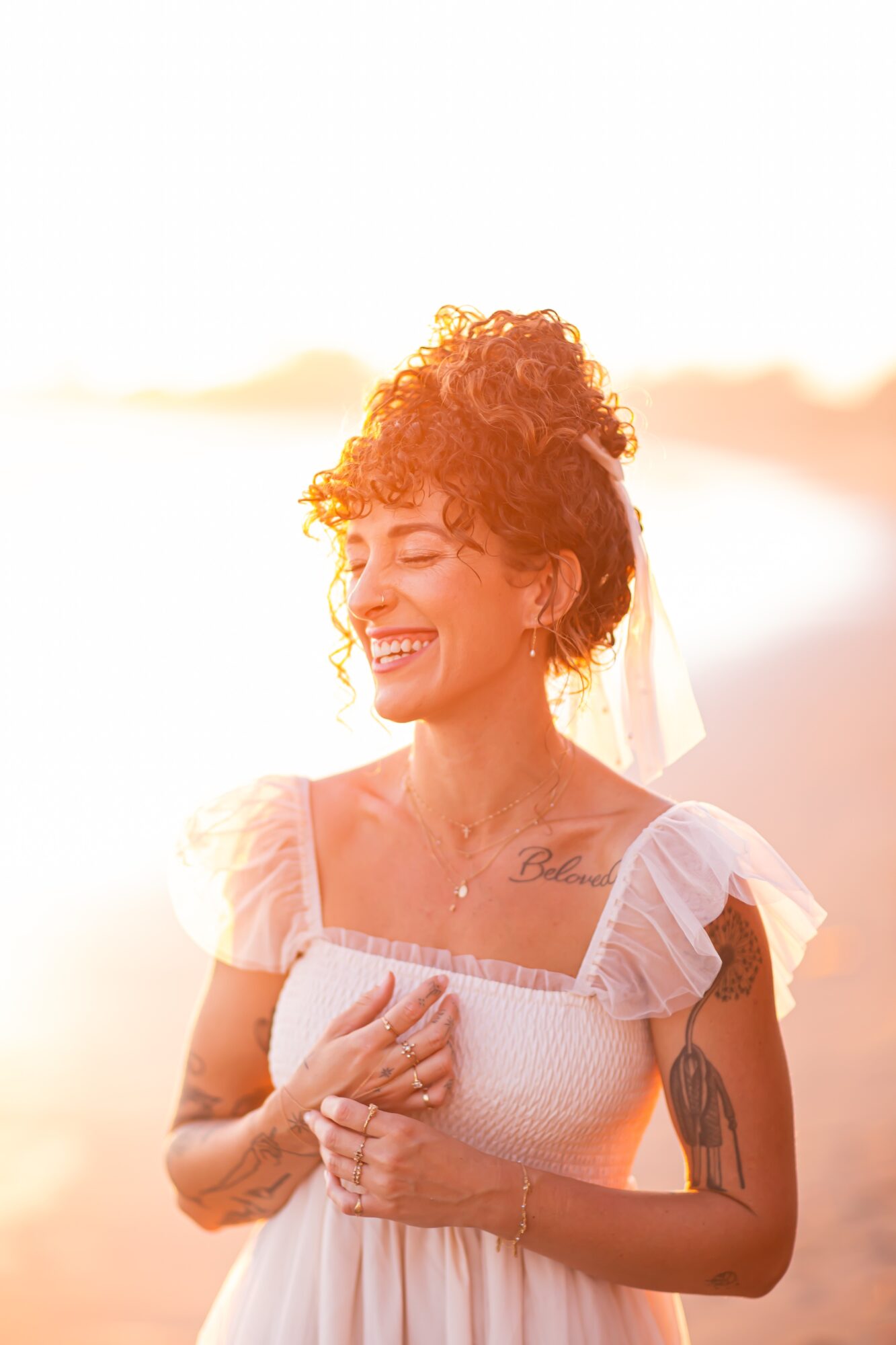 Woman with curly hair smiling outdoors, wearing a white dress with puffed sleeves, holding her chest, bright background