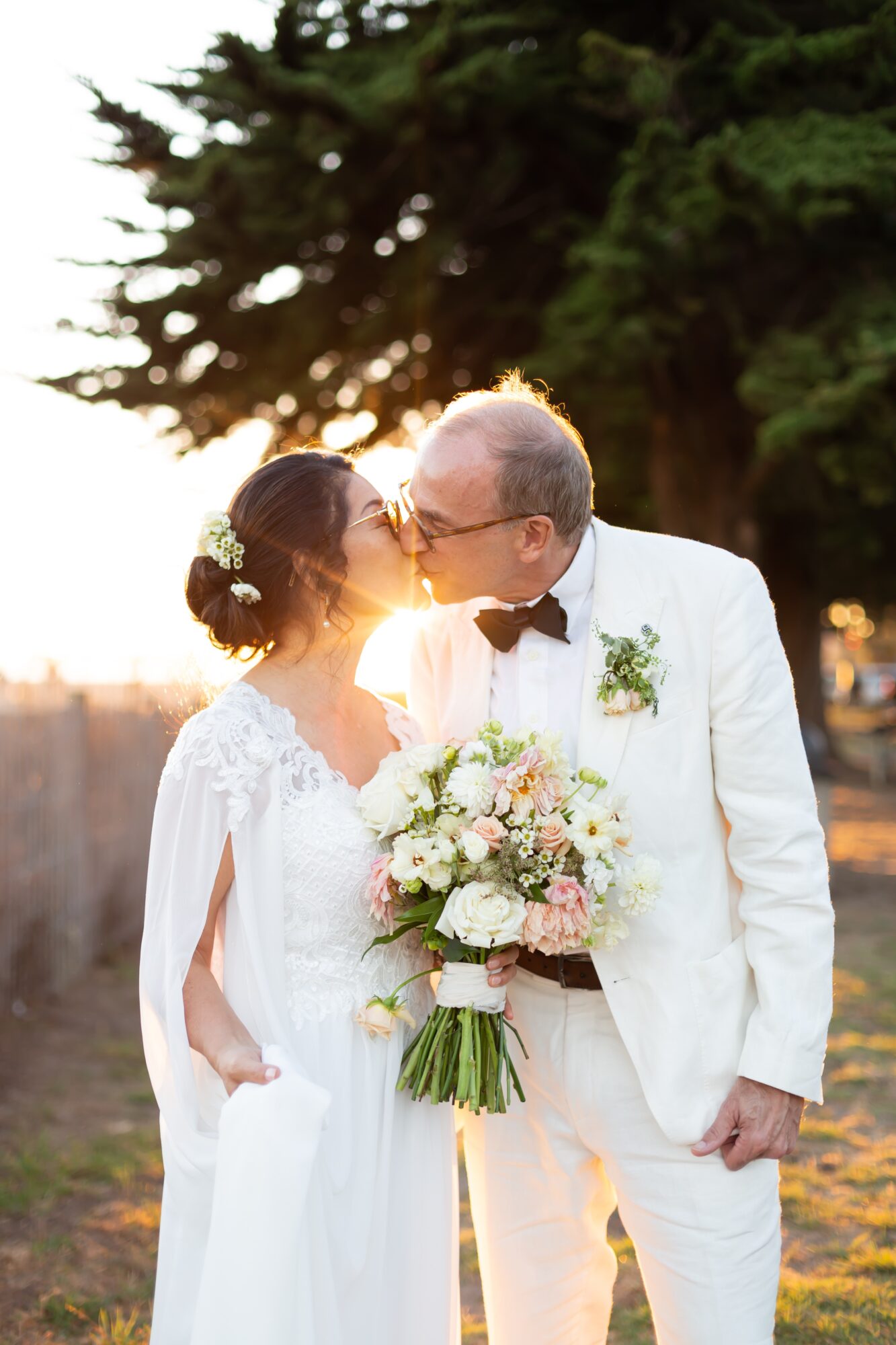 A bride and groom kiss outdoors during sunset, with trees in the background, the bride holding a bouquet of flowers.