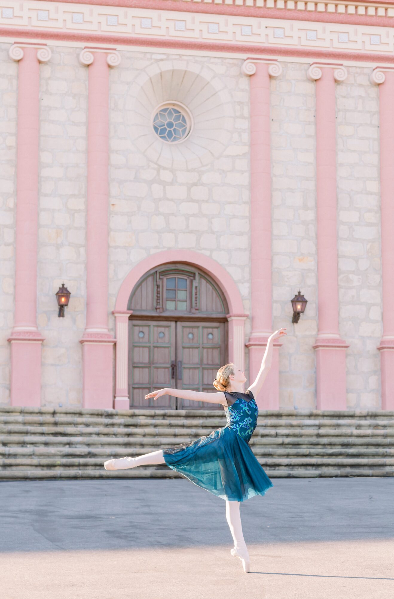 Ballet dancer in blue dress performs on steps in front of a pink and white building with columns.