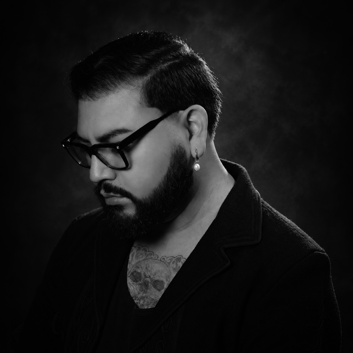A man with glasses and a beard looks downward, wearing a dark jacket against a dark background.