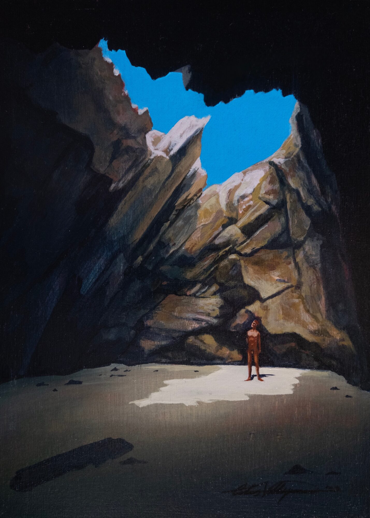A person standing on a sandy beach between large rock formations with a bright blue sky visible through a gap.