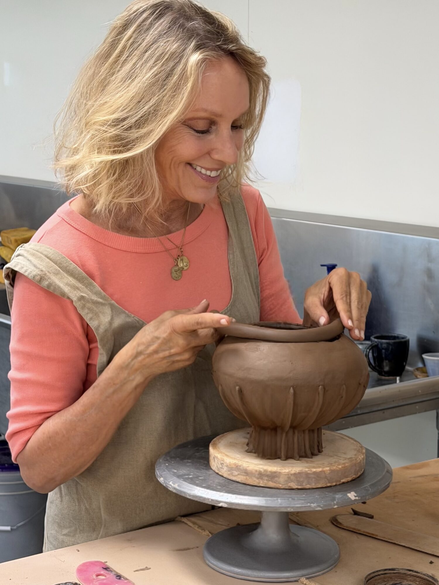 Woman smiling while shaping a clay pot in a pottery studio, wearing a pink shirt and beige apron, with pottery tools nearby.