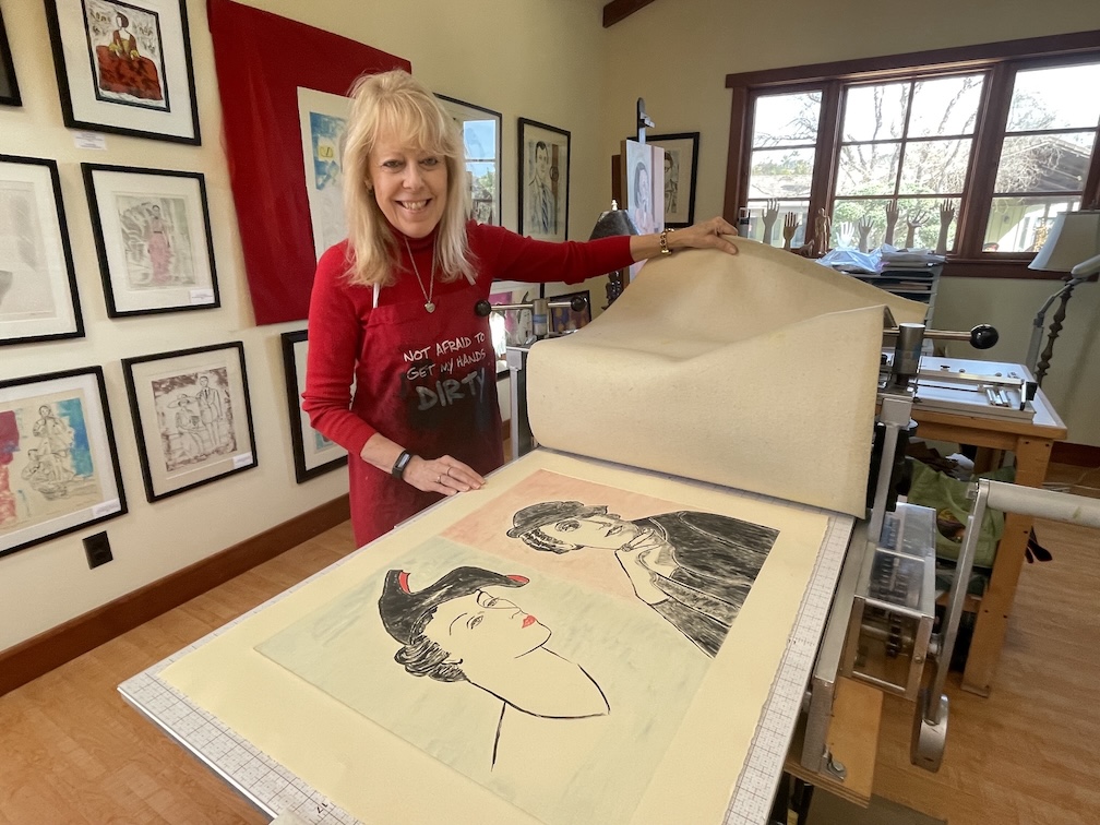 Woman with blonde hair in red sweater smiling at large art book with drawings, in a room with framed artwork and large windows.