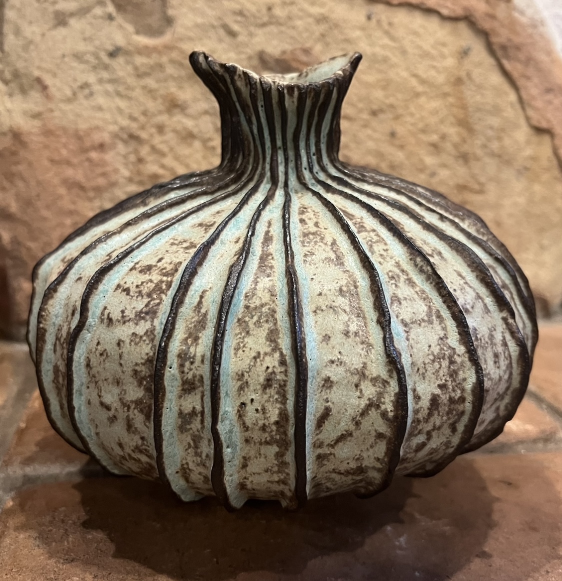 Decorative ceramic vase with vertical black lines and a textured surface, placed on a wooden surface against a brick wall.
