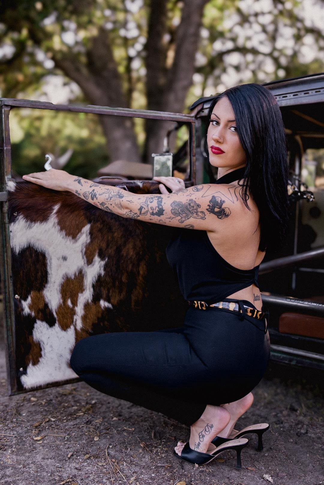 Woman with black hair and tattoos squats near a cow, outdoors with trees in background.