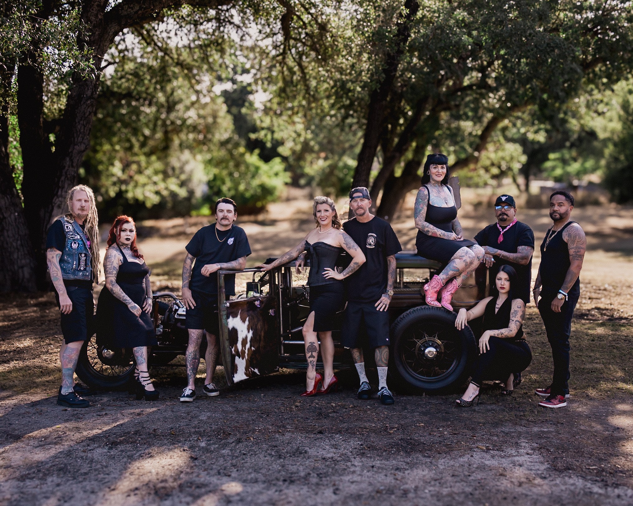 Group of nine people outdoors with trees, standing and sitting on a vehicle and tire, in sunlight.