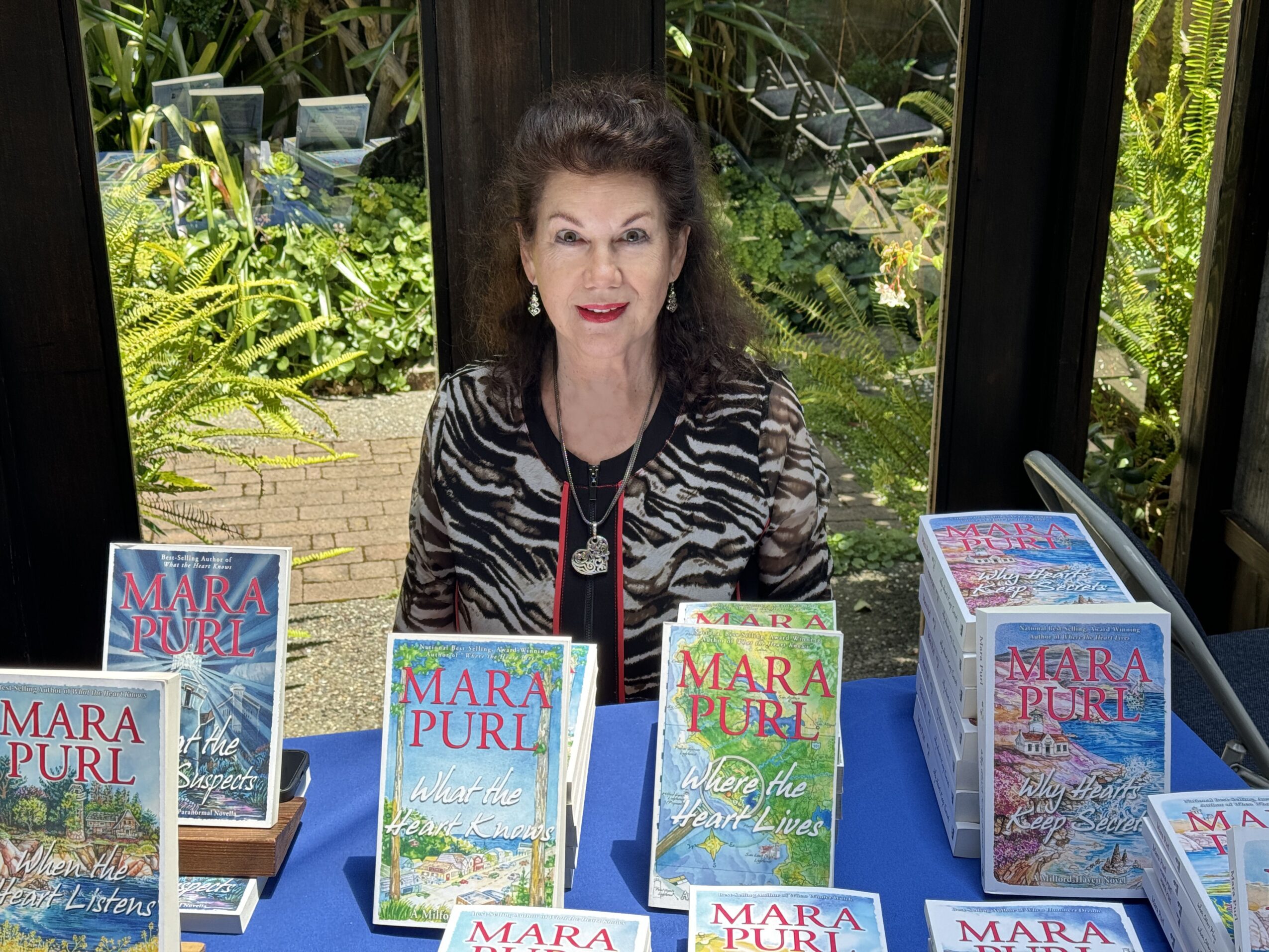 Woman sitting at a table with books, outdoors with greenery behind glass windows.