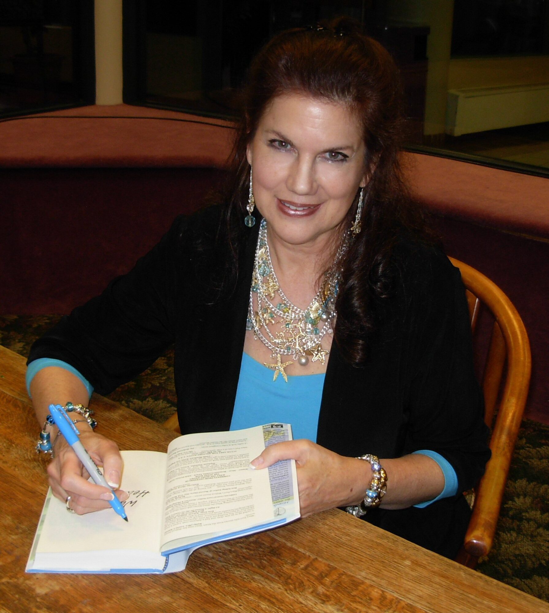 Woman with dark hair, wearing jewelry and a black jacket, sitting at a table with an open book and pen.