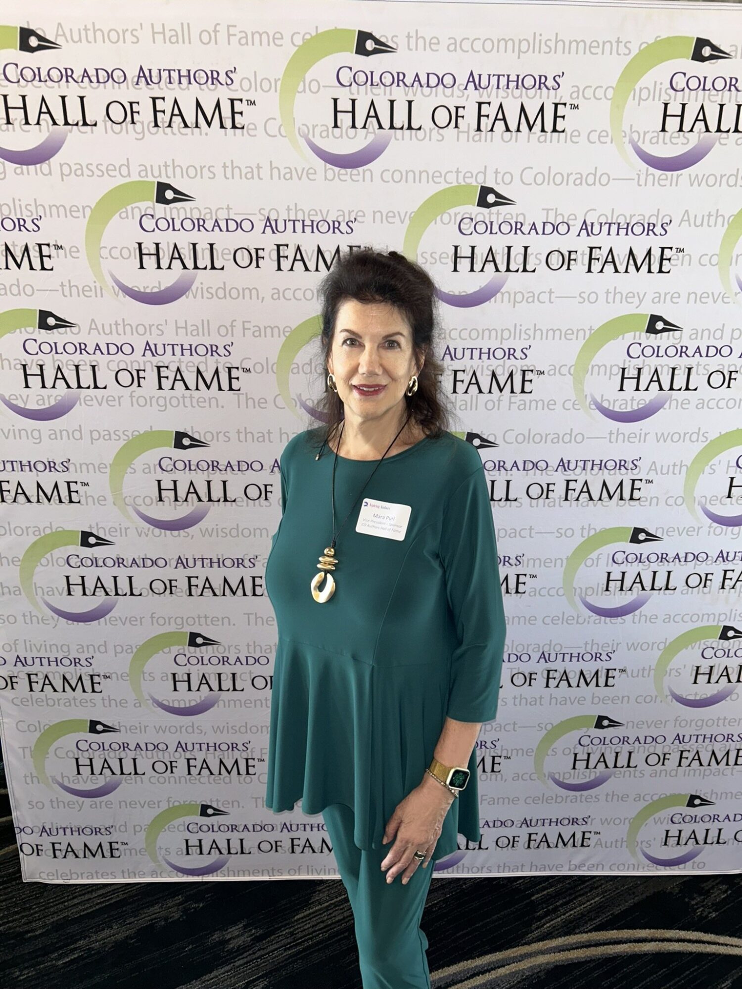 Woman with dark hair in green dress standing in front of Colorado Authors' Hall of Fame backdrop, smiling, wearing a necklace and badge.