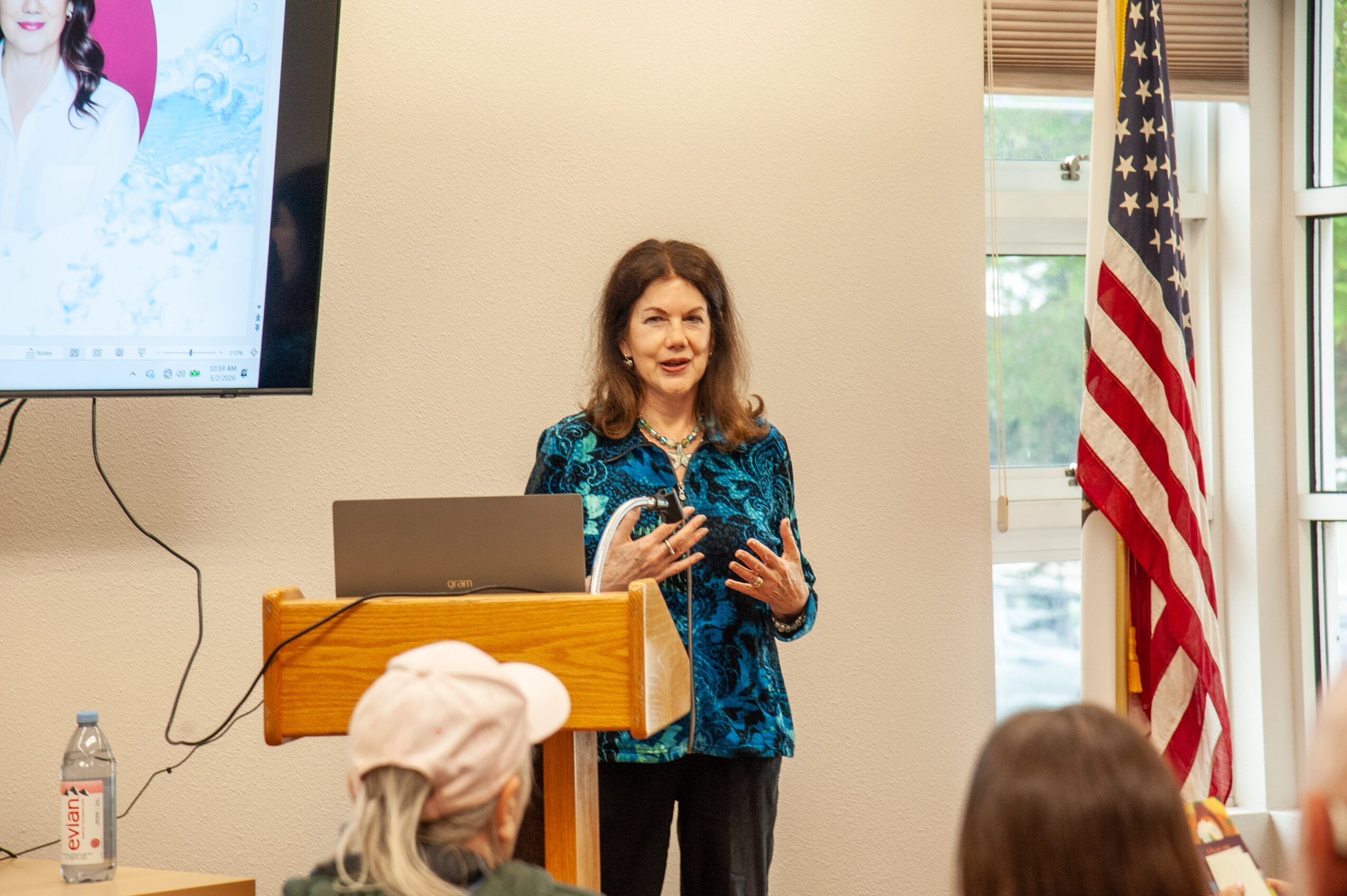 Woman speaking in front of an audience, with a laptop on a wooden podium, American flag nearby, and a screen on the wall.