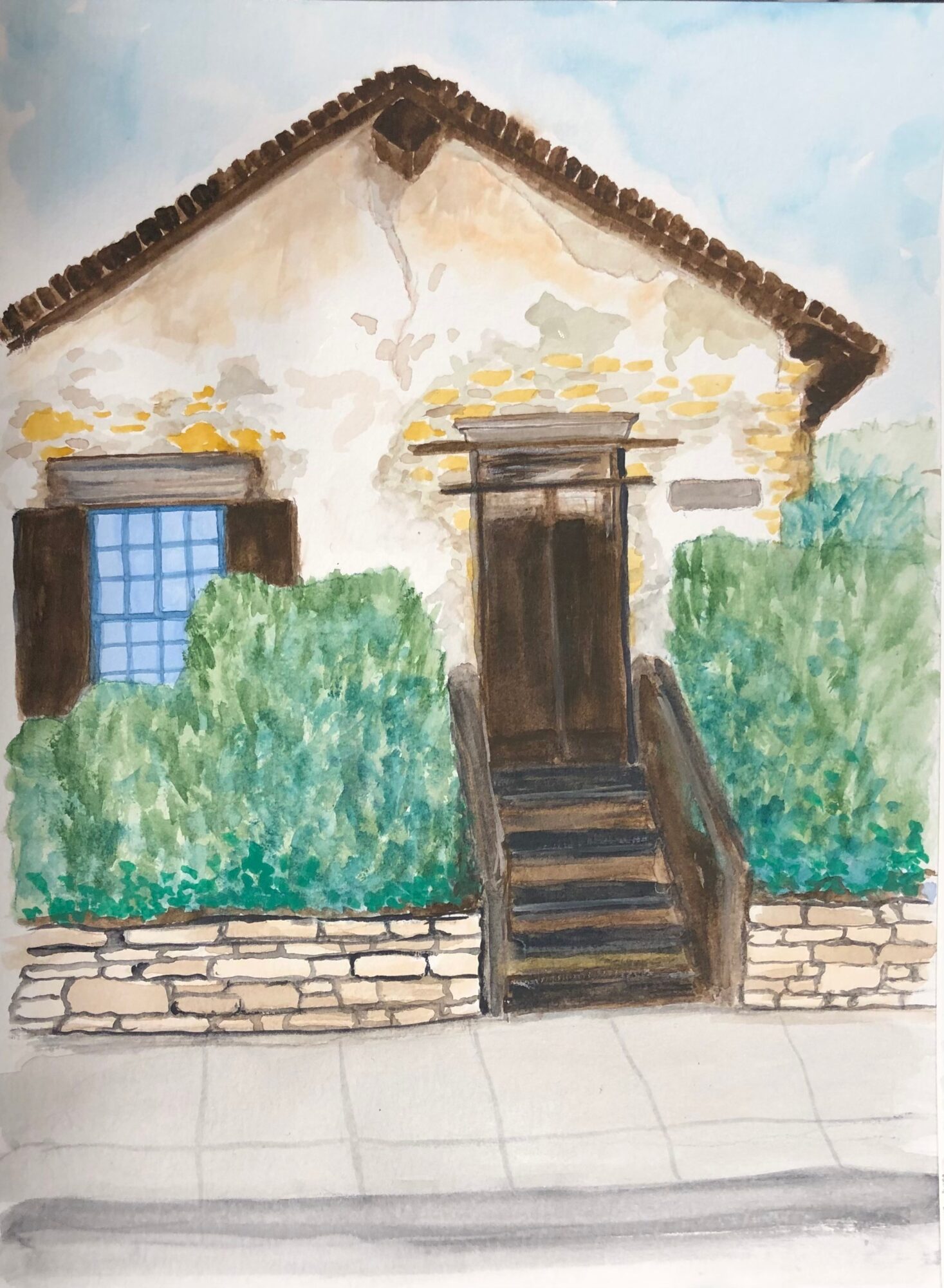 Small house with a steep roof, wooden stairs, surrounded by green bushes, and a stone wall in front.