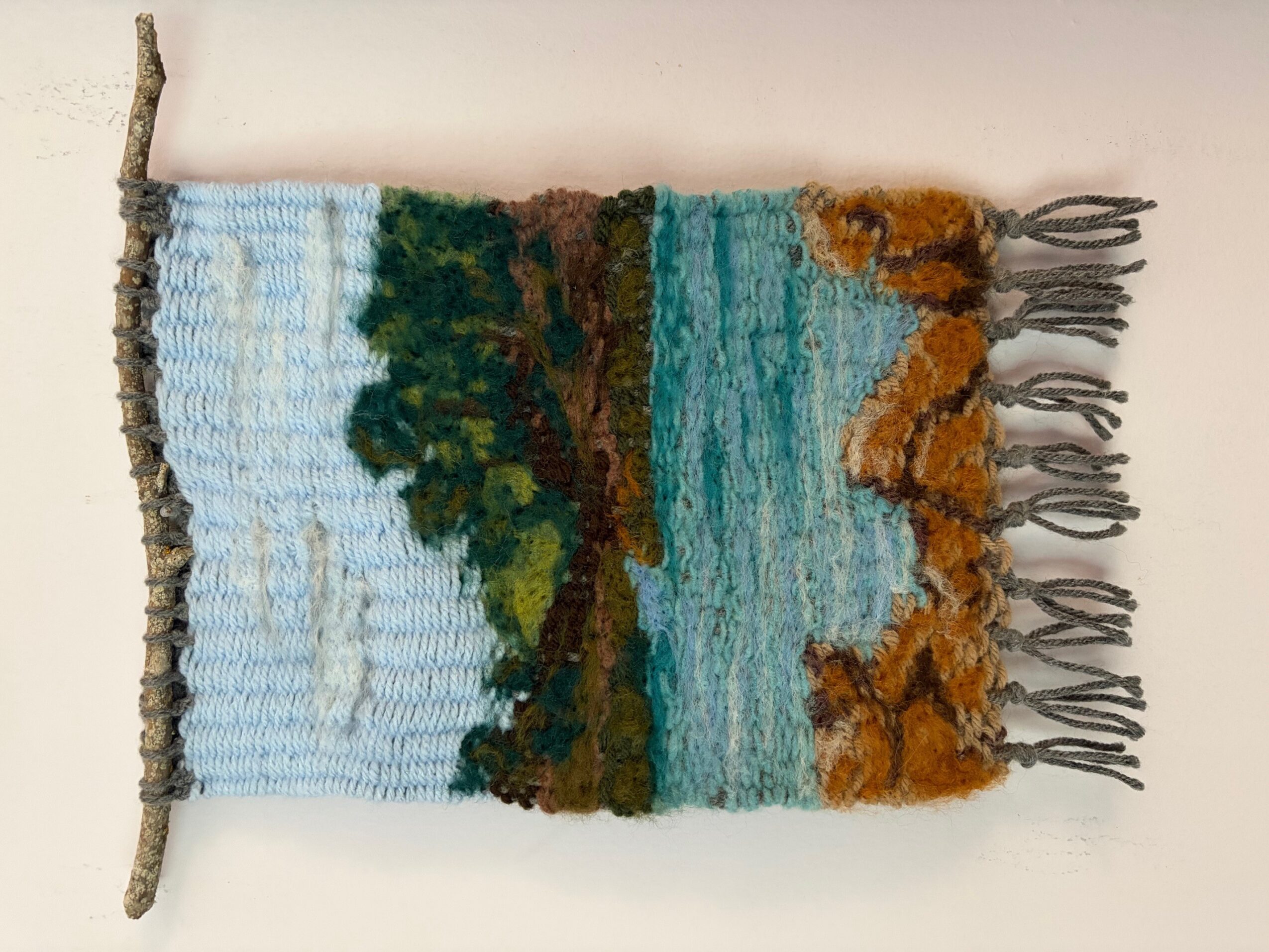 Textured woven wall hanging with landscape scene, trees, water, and orange foliage, hanging on a white wall.