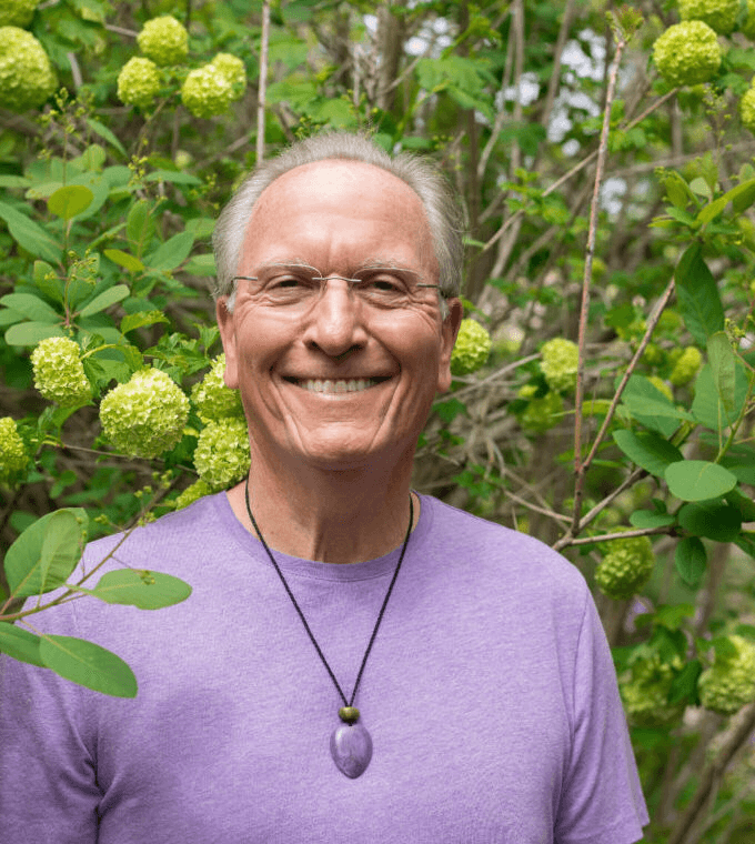 Smiling elderly man with glasses and gray hair outdoors, wearing a purple shirt and a purple pendant necklace, surrounded by green foliage and yellow-green flowers.