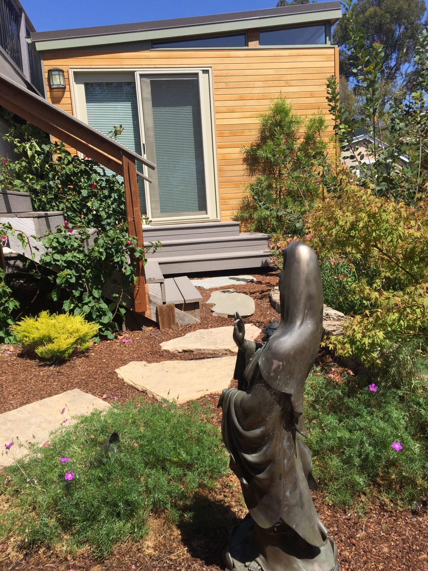 Garden with stone pathway, greenery, and a wooden house with a glass door in the background.