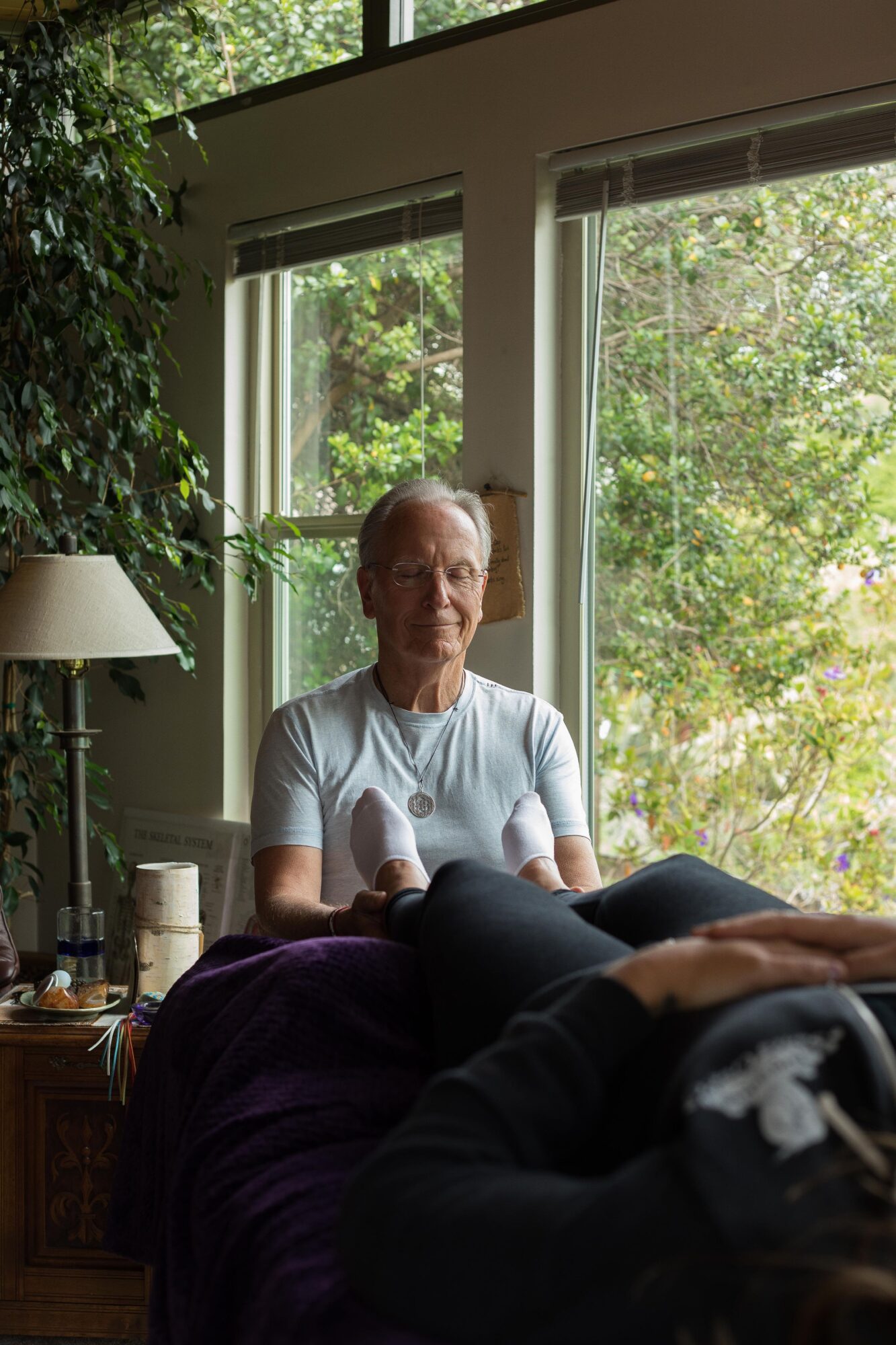 Elderly person sitting on a bed near large windows with greenery outside, sunlight streaming in, holding a book or paper.