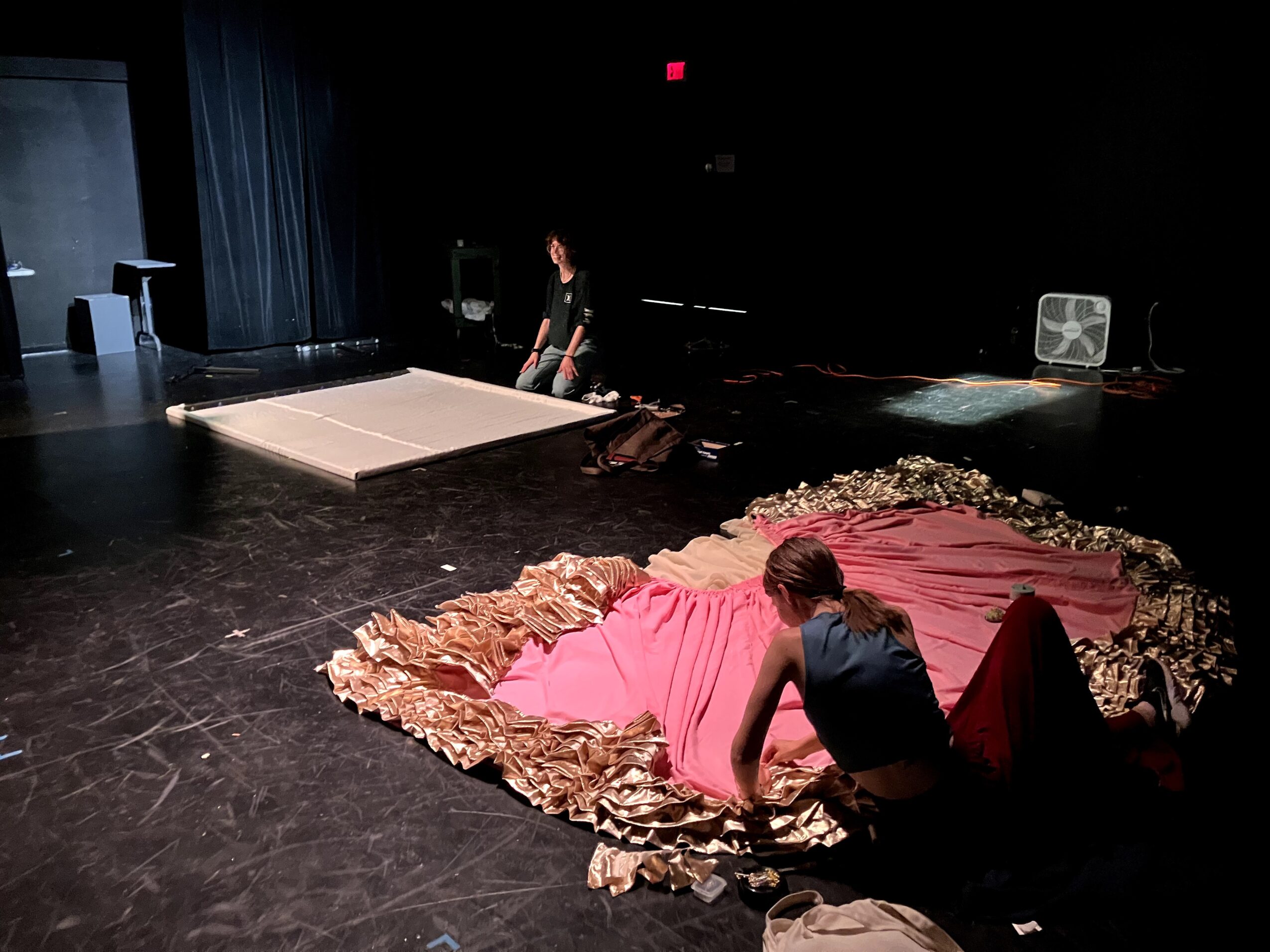 Two people prepare a stage with fabric and decorative elements, one kneeling and one standing, in a dark theater setting.