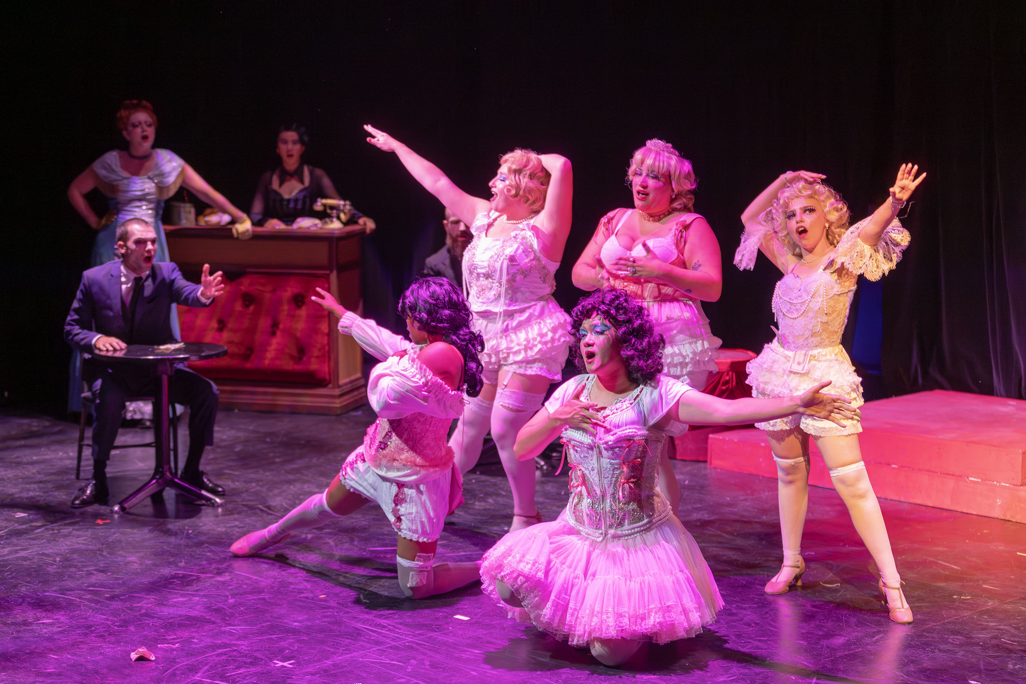 Group of performers on stage, some dancing and others playing instruments, with a dark background and stage lighting.