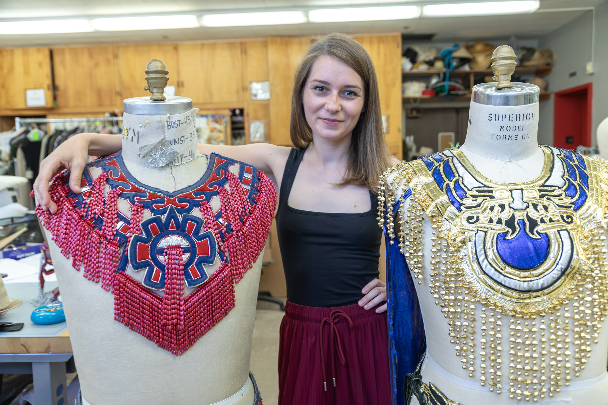 Young woman standing between two mannequin displays of elaborate jewelry, in a workshop setting.