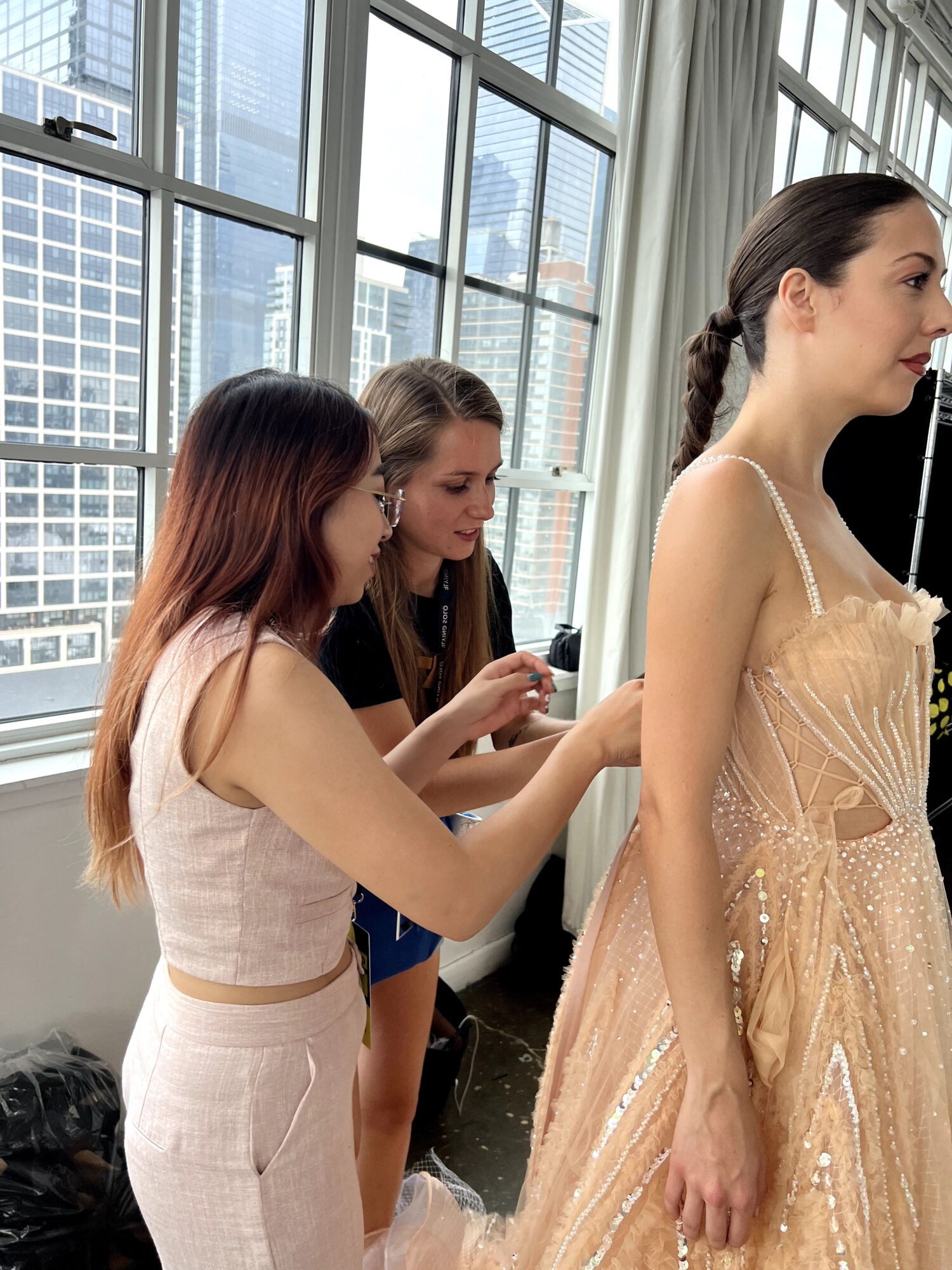 Three women in a bright room with large windows, one in a beige dress, two using a phone.