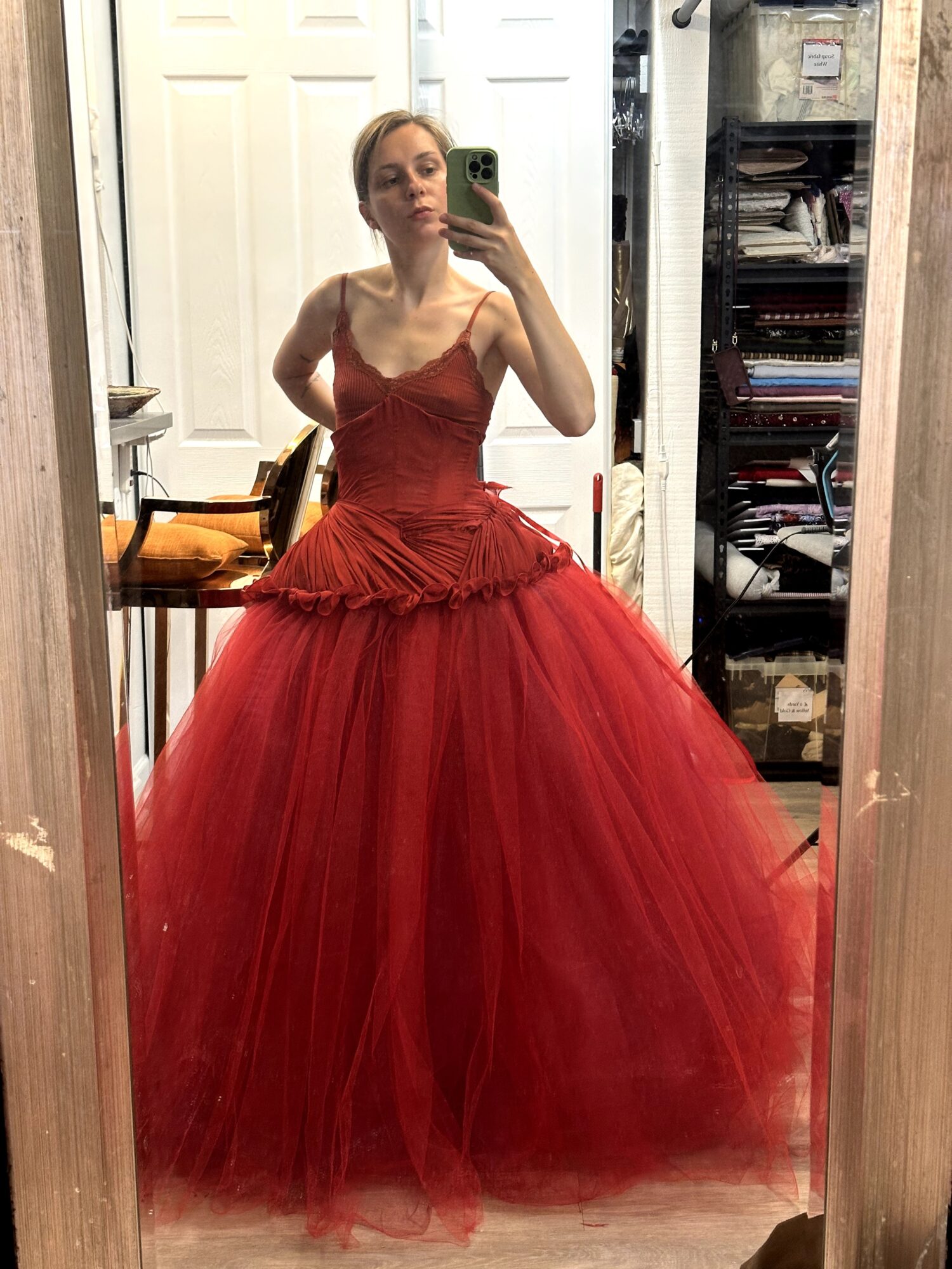 Person in a red gown taking a mirror selfie in a room with shelves and a door.