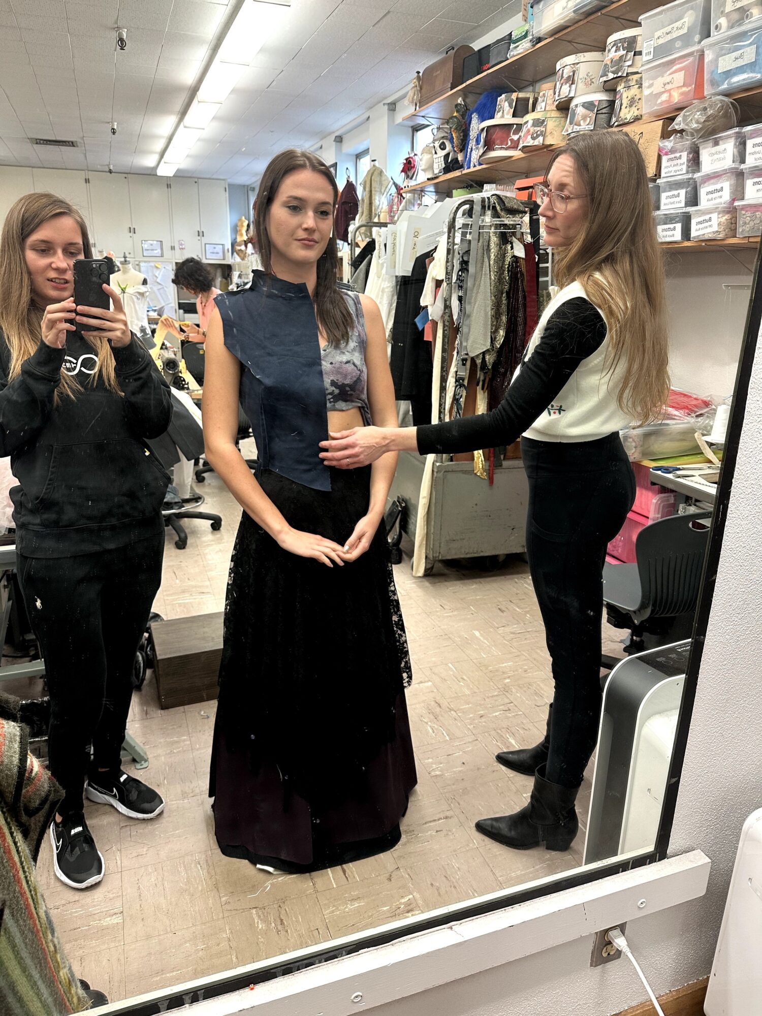 Three women in a clothing store, one trying on a dress, others assisting and taking a photo, shelves with boxes and clothes behind.