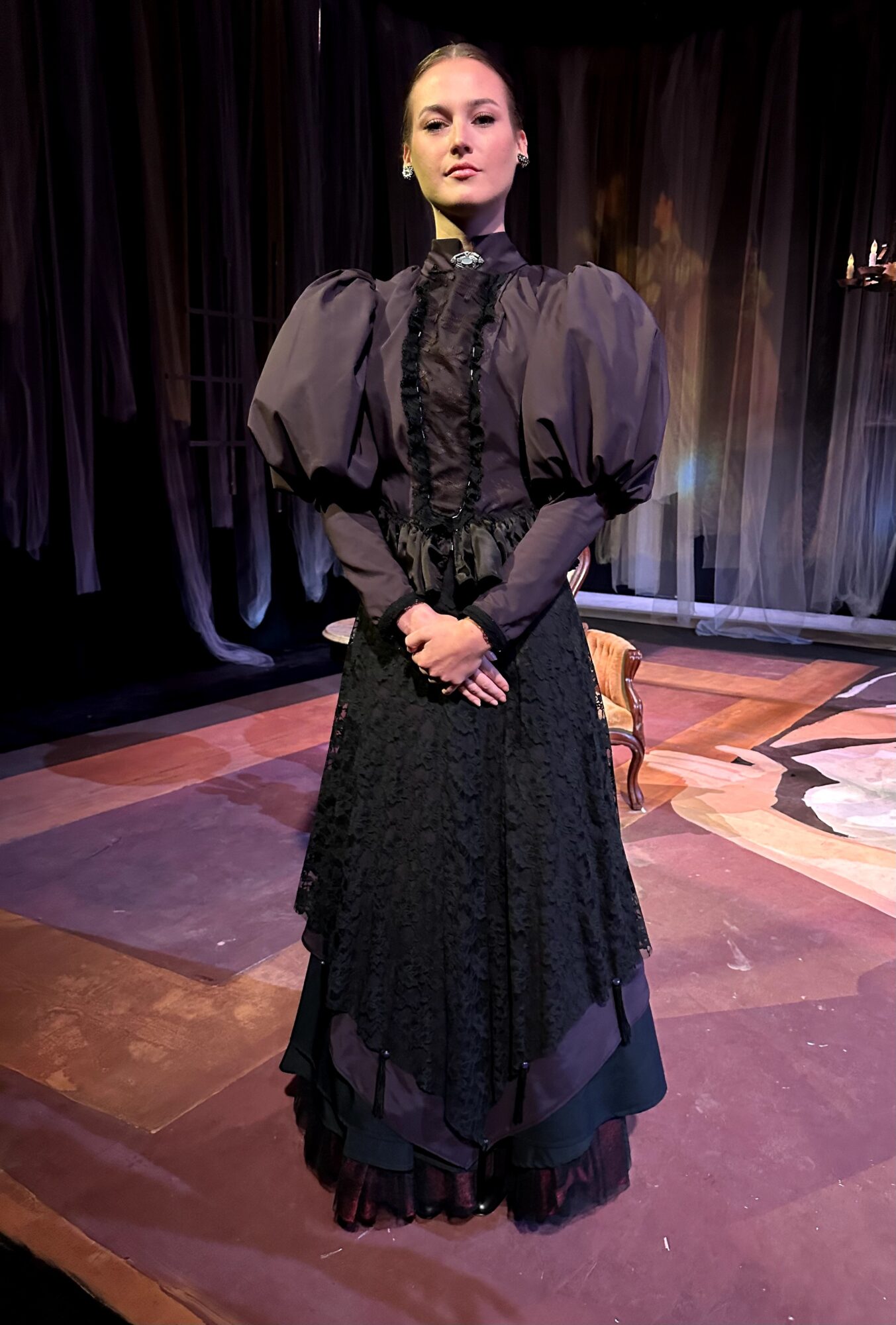 Woman in black dress with puffed sleeves standing on stage with dark curtains behind her.
