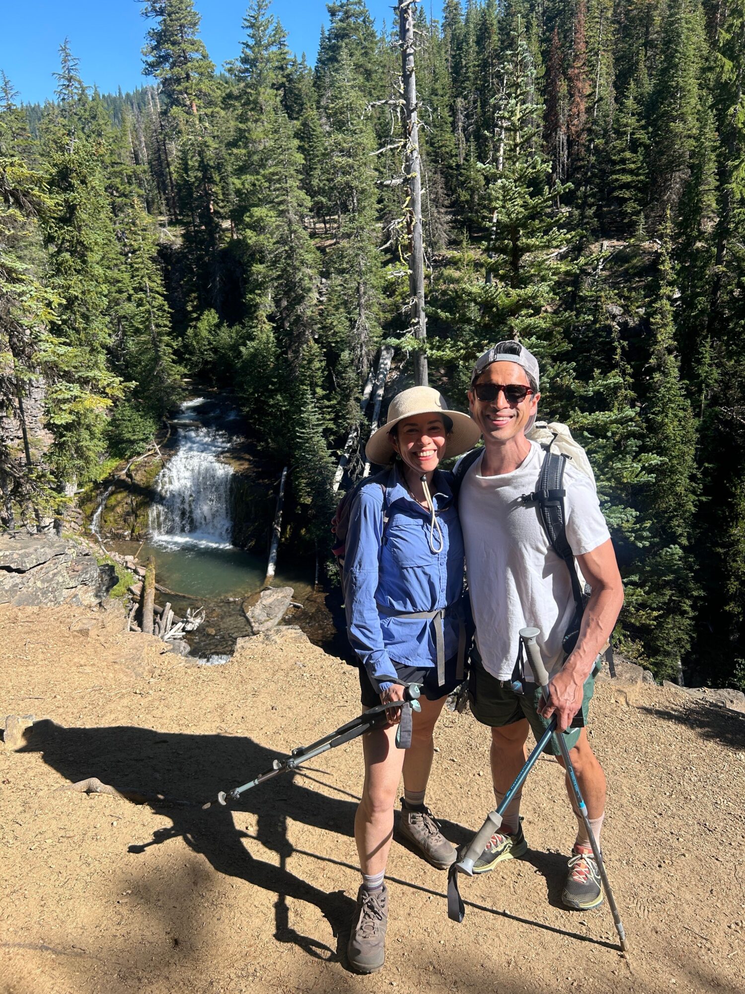 Two hikers with backpacks and walking sticks stand on a dirt trail near a small waterfall surrounded by trees.
