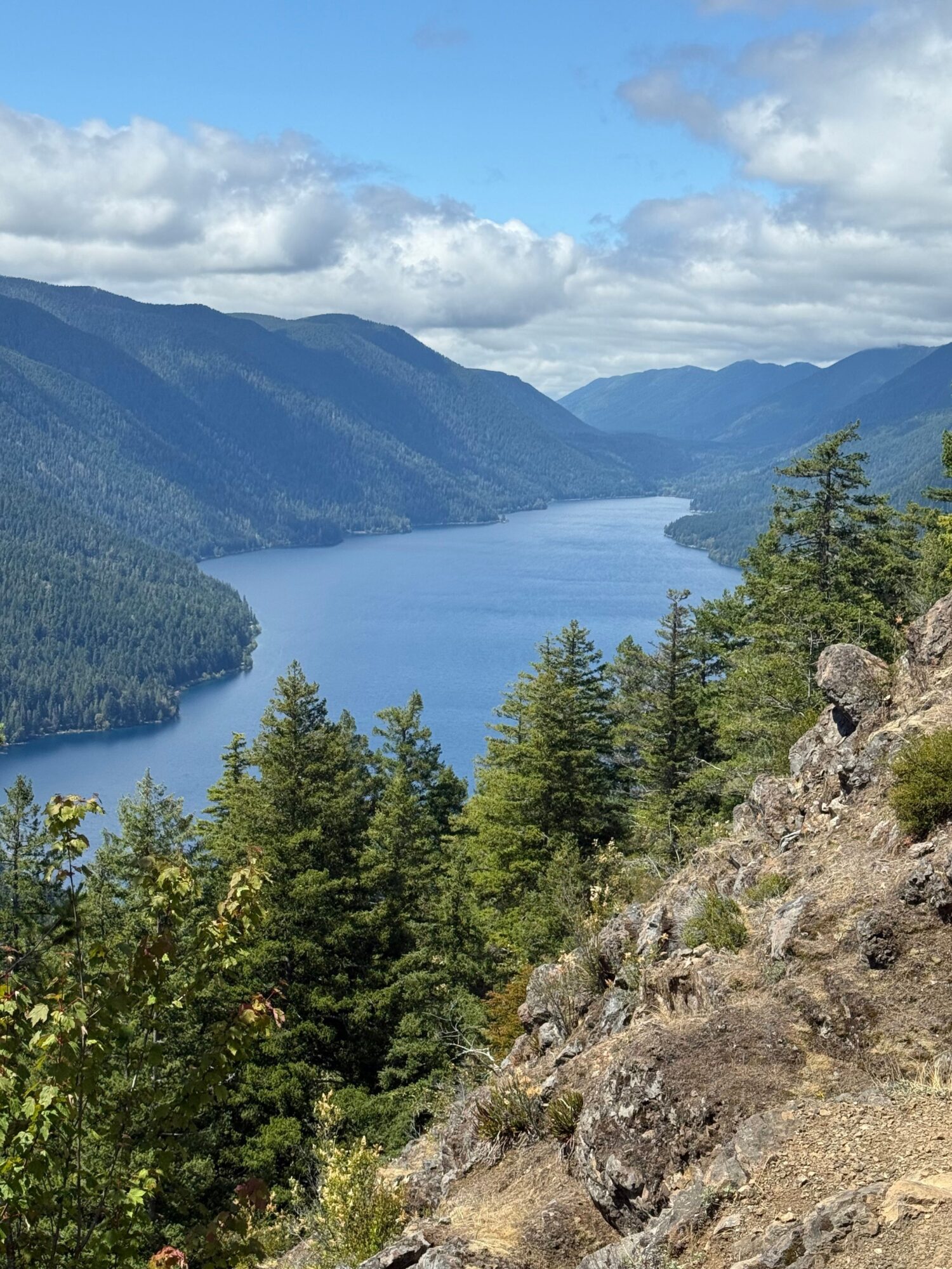 Mountain landscape with trees, rocky terrain, and a cloudy sky, viewed from an angle.
