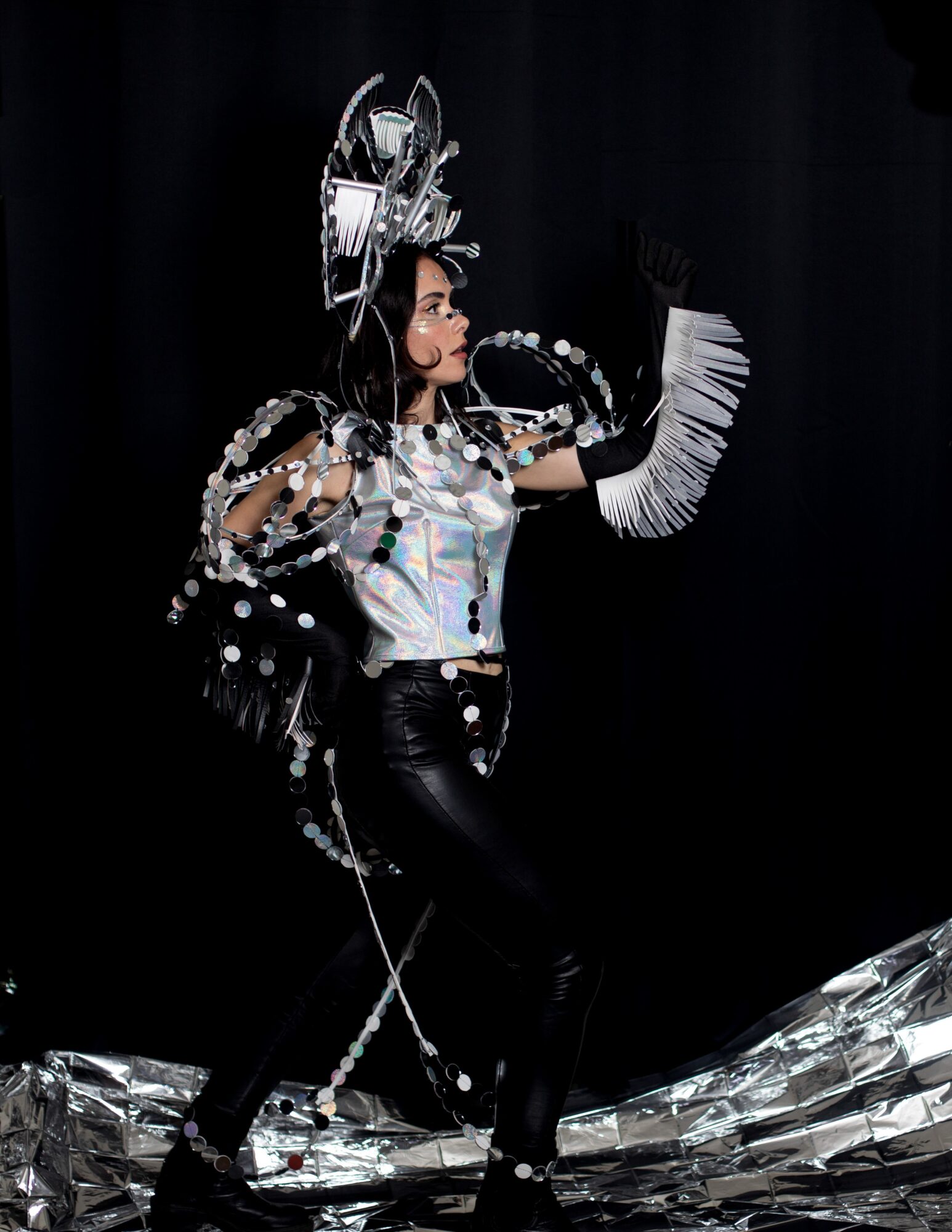 Person in elaborate costume with metallic and feathered accessories, standing on a shiny surface against a dark background.