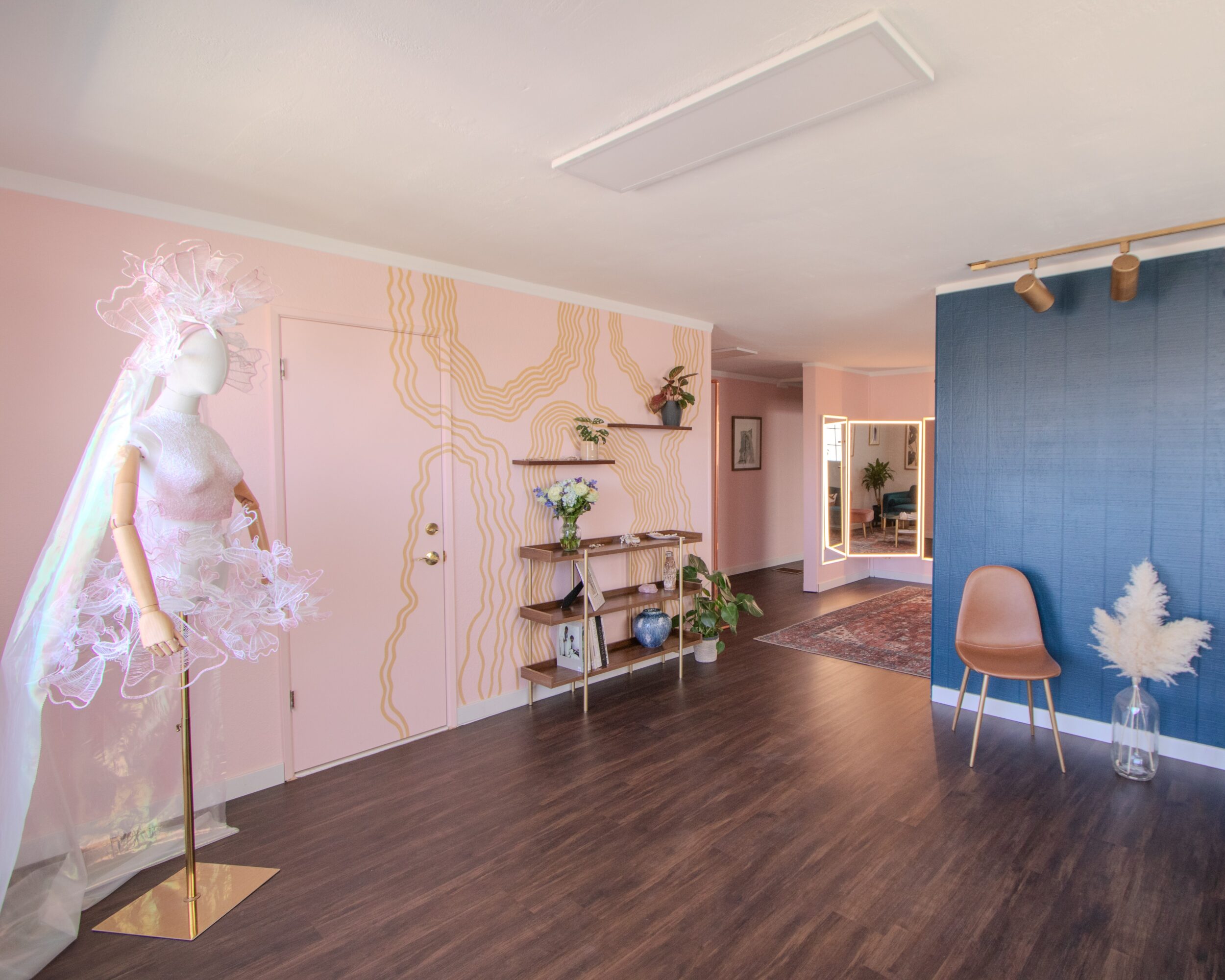 Interior room with a mannequin, shelves, plants, and chairs, featuring pink and blue walls, and wooden flooring.