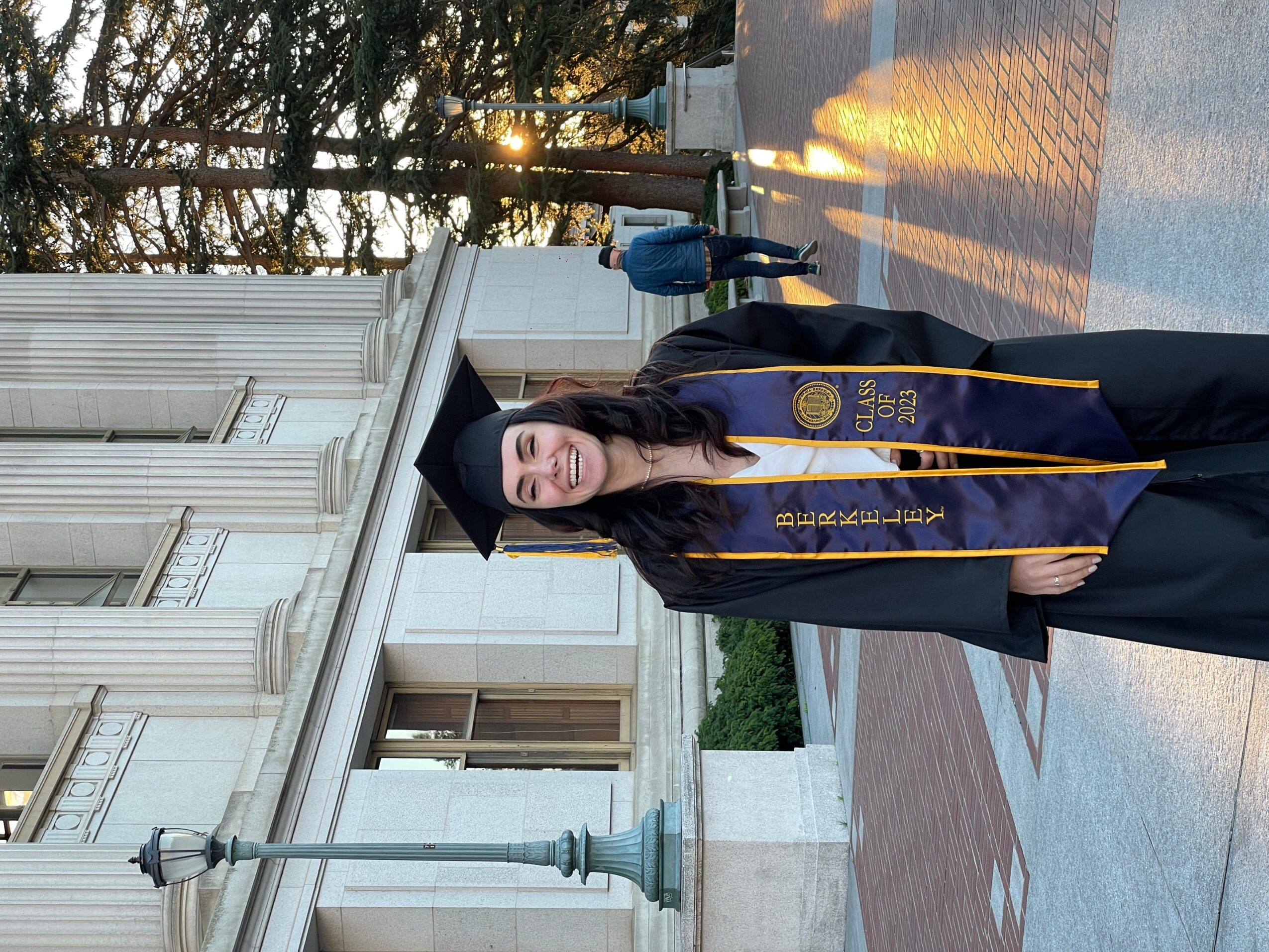 Graduate in cap and gown standing outdoors with building in background, sunlight, and trees.
