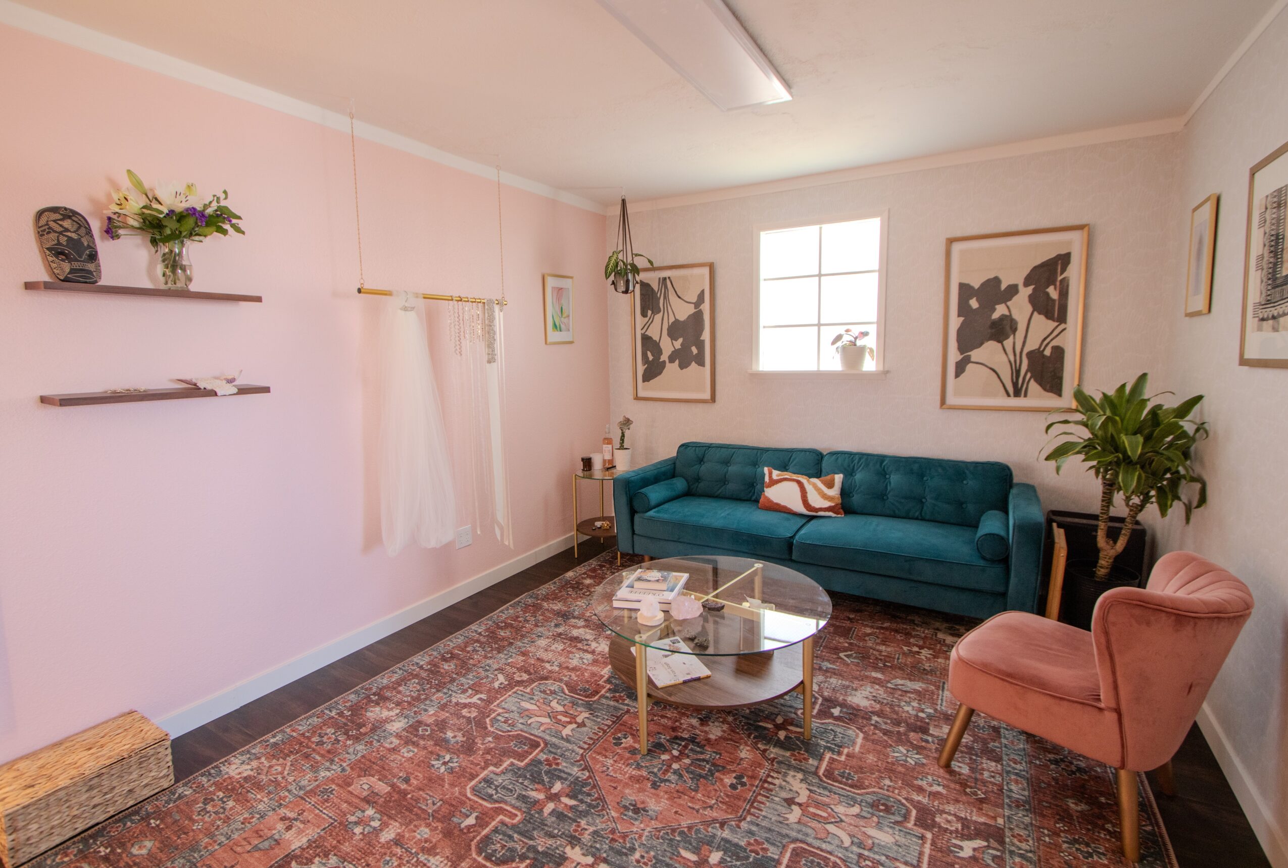Living room with pink walls, teal sofa, pink armchair, round coffee table, and patterned rug, with wall art and plants.