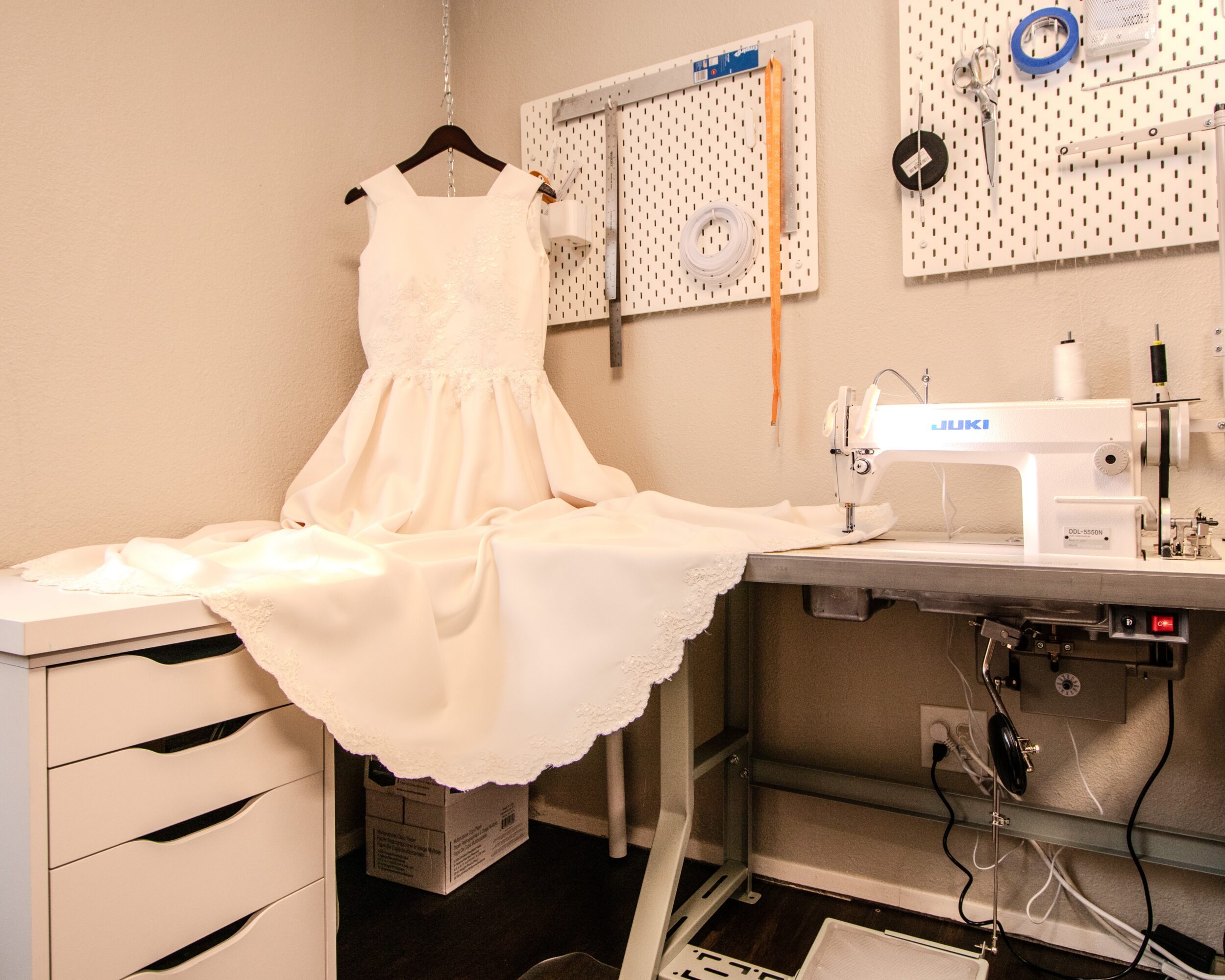 A white dress hangs on a black hanger above a sewing machine in a room with sewing tools on a pegboard.