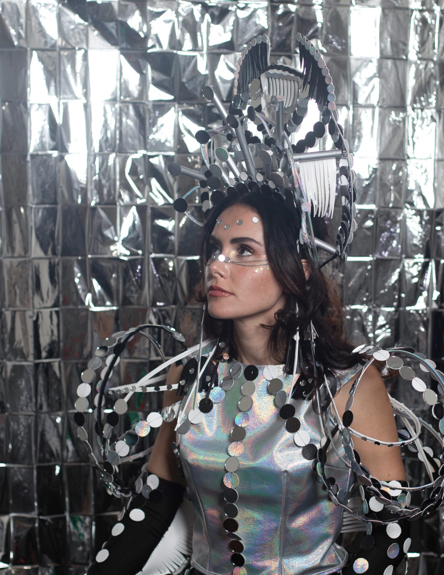 Woman with glasses and dark hair in futuristic metallic costume with reflective background, looking to the side.