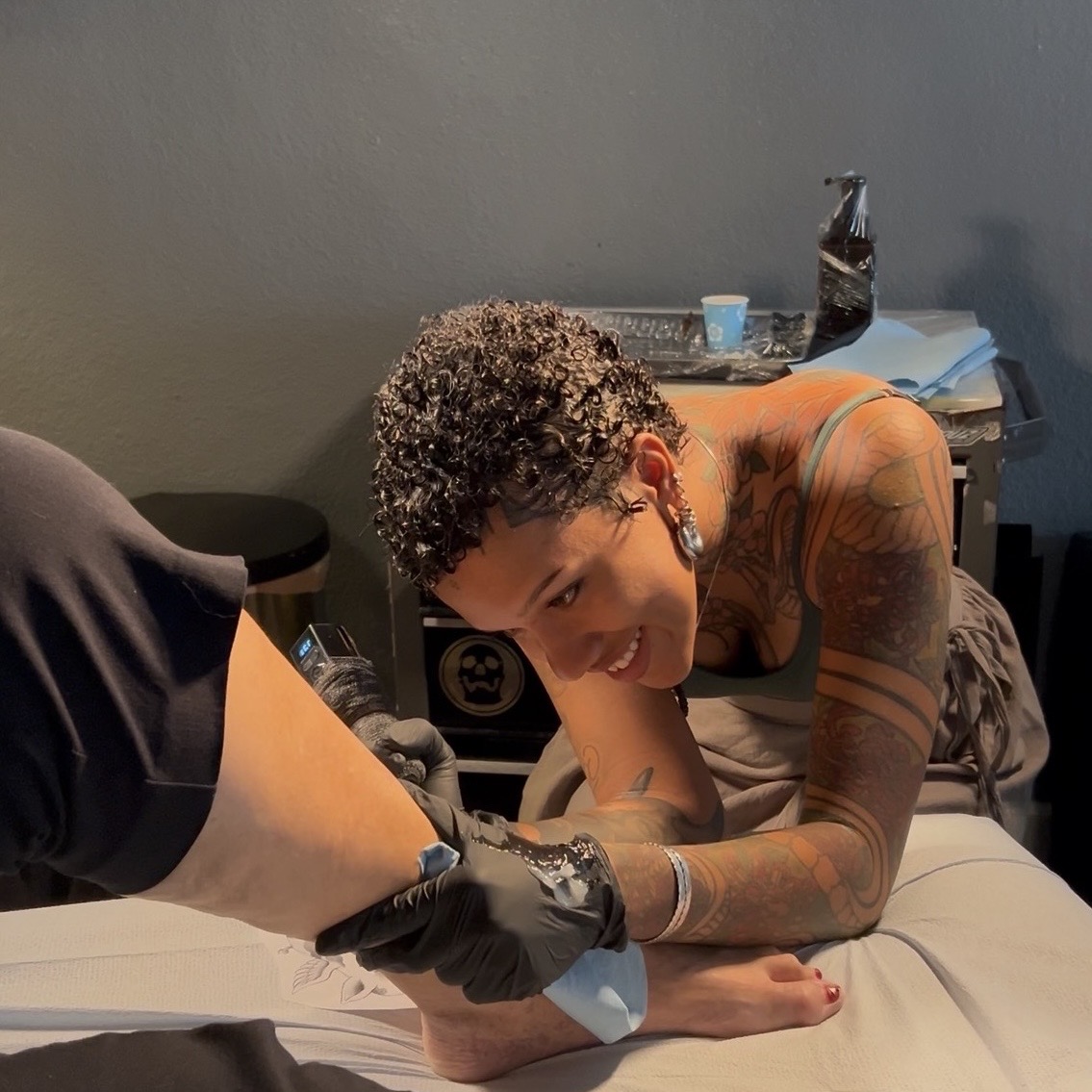 Person with curly hair and tattoos lying on a bed, receiving a tattoo from an artist wearing black gloves.